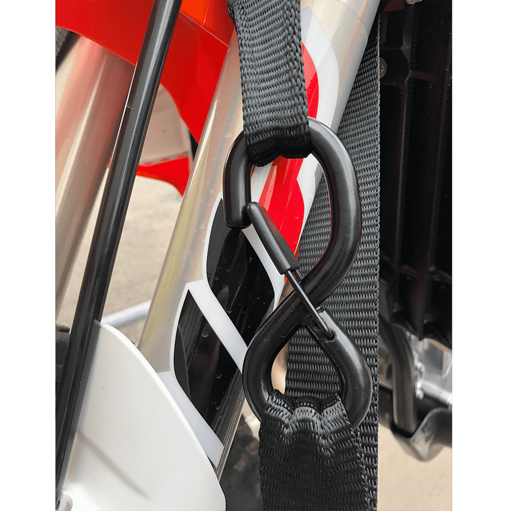 Nahaufnahme eines O'NEAL DE LUXE Zurrgurte LOGO aus schwarzem Nylon mit einem Kunststoff-S-Haken, der an der Vordergabel eines Motorrads befestigt ist, mit Soft Hook Tie Downs und roten, schwarzen und weißen Details im Hintergrund.