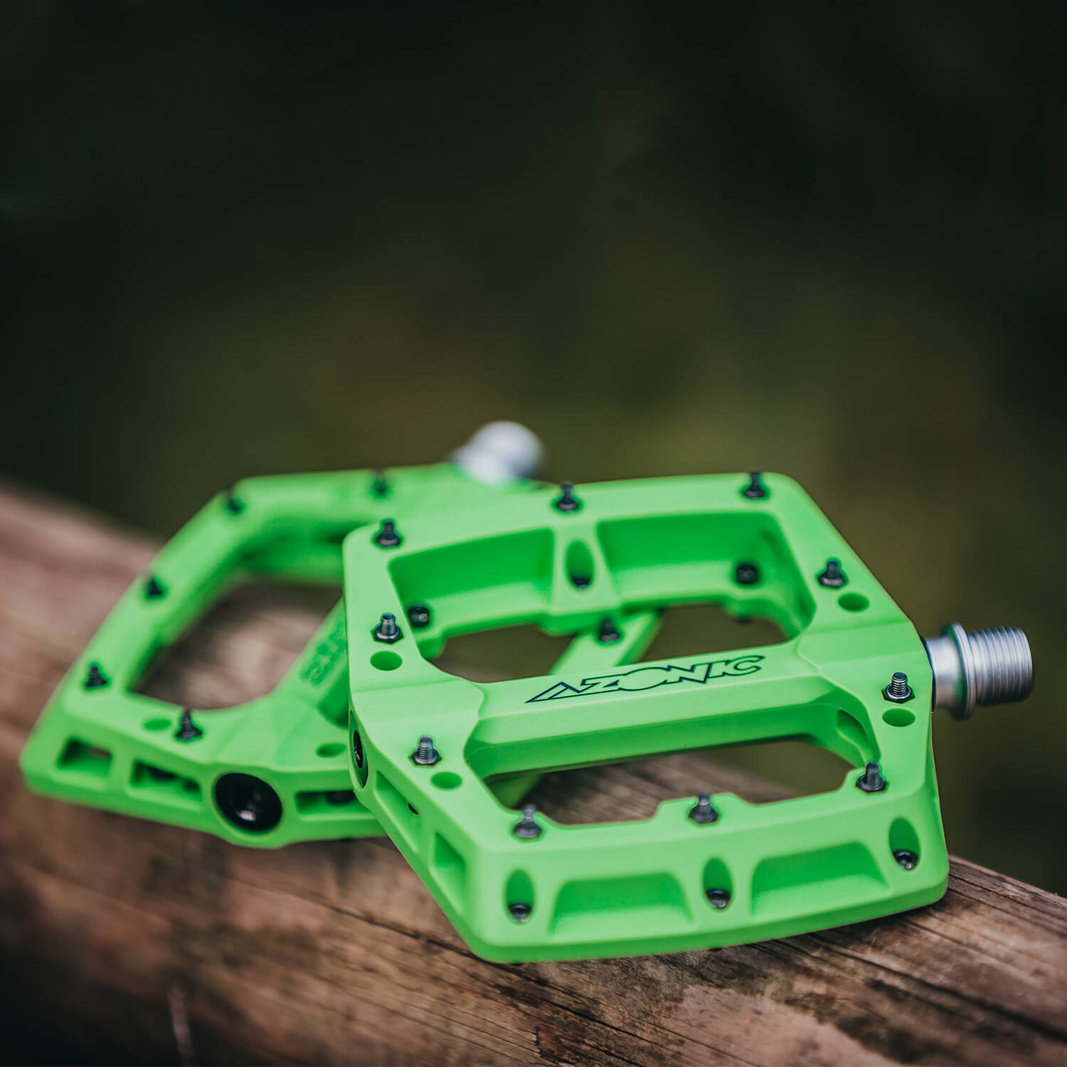 Eine Nahaufnahme von zwei grünen Azonic-Fahrradpedalen, die auf einer Holzoberfläche ruhen. Die Pedale sind robust gebaut und verfügen über mehrere Stifte für den Halt sowie eine Metallspindel zur Befestigung am Fahrrad. Der Hintergrund ist verschwommen und scheint eine Außenkulisse abzubilden.