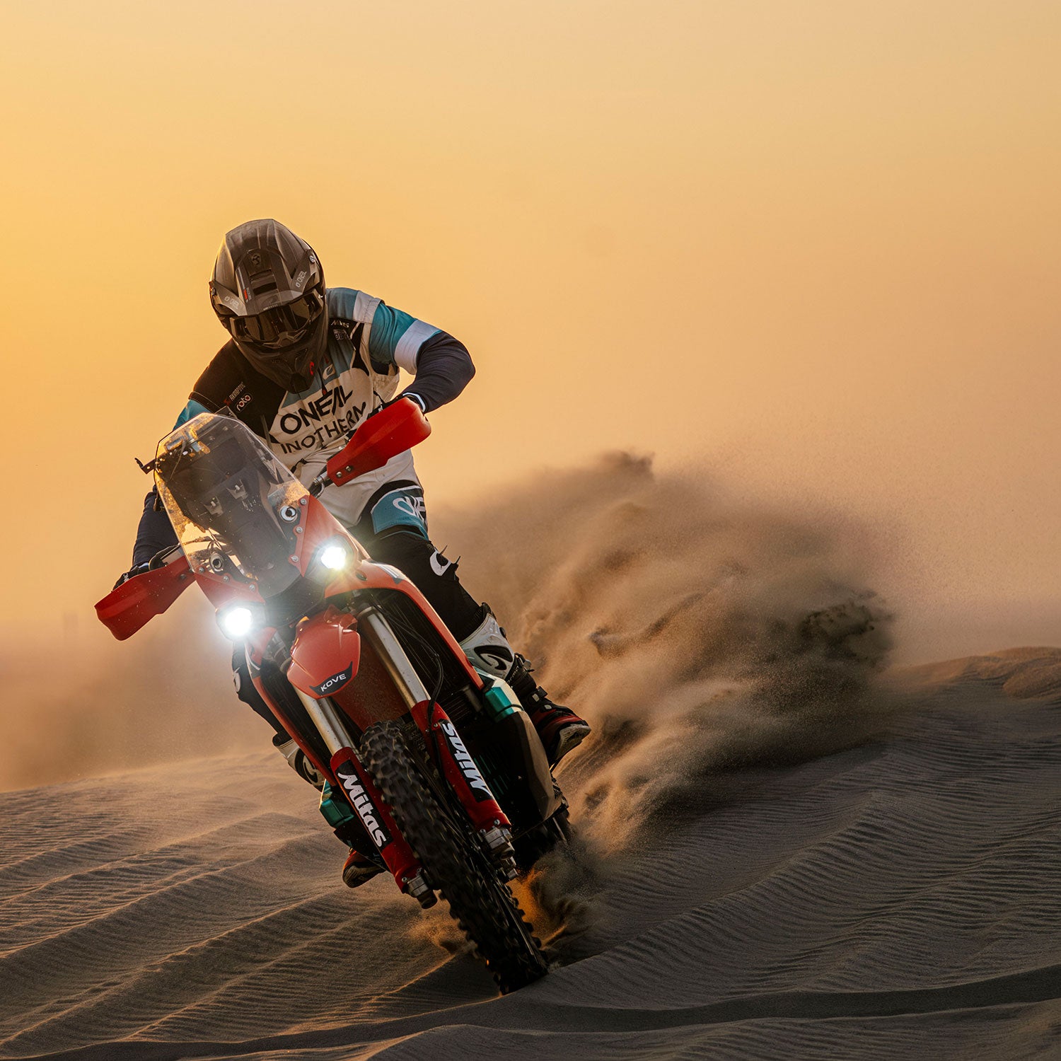 Ein Motorradfahrer fährt bei Sonnenuntergang mit hoher Geschwindigkeit über sandige Dünen und wirbelt dabei Sandwolken auf. Die Scheinwerfer des Motorrads sind eingeschaltet, und der Fahrer trägt einen Helm und Schutzkleidung.