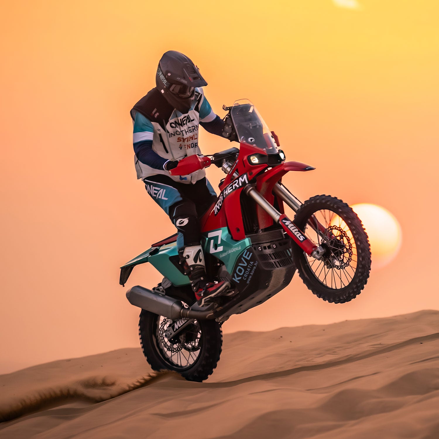 Ein Motorradfahrer in voller Montur vollführt einen Wheelie auf einem rot-grünen Dirt Bike über sandige Dünen bei Sonnenuntergang, während die Sonne tief steht und im Hintergrund leuchtet.