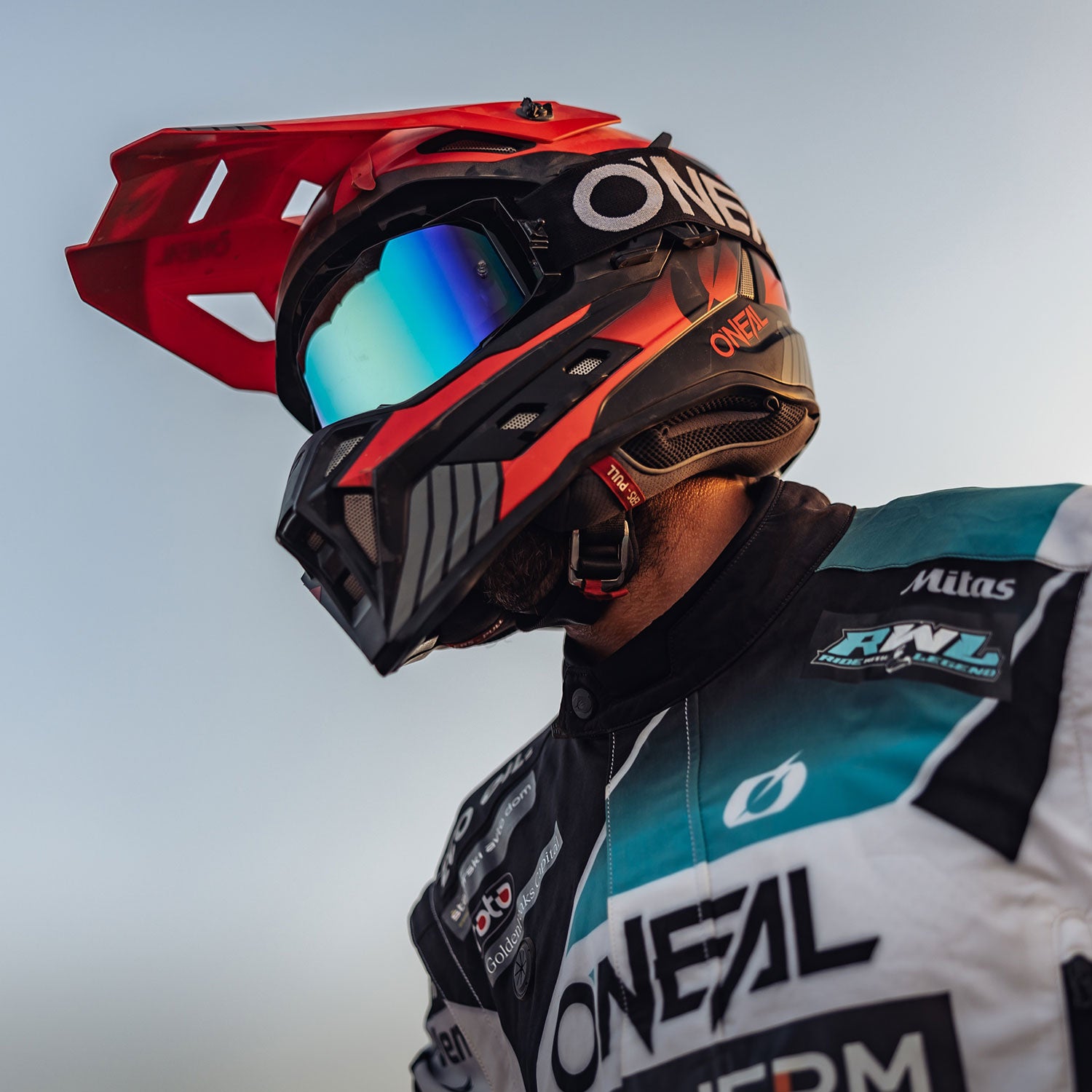 Ein Motocross-Fahrer mit einem schwarz-roten Helm mit reflektierender Schutzbrille und einem weißen Rennanzug mit verschiedenen Logos, fotografiert aus einem niedrigen Winkel vor einem klaren Himmel.