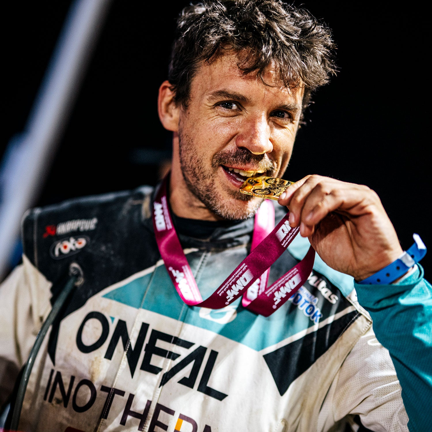 Ein Mann im Rennanzug trägt eine Goldmedaille, die an einem kastanienbraunen Band um seinen Hals hängt, lächelnd und mit triumphierendem Blick. Der Hintergrund ist dunkel und unscharf.