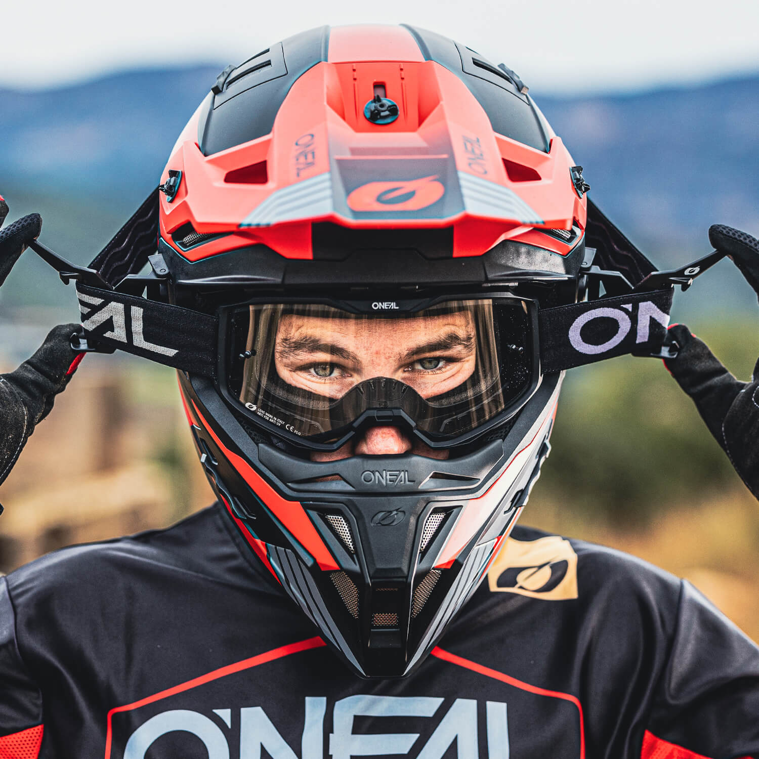 Eine Person mit einem schwarz-roten O'Neal-Motorradhelm rückt ihre Schutzbrille zurecht. Die Person trägt passende O'Neal-Ausrüstung und Handschuhe an beiden Händen. Der Hintergrund ist verschwommen und hebt den Helm und die Ausrüstung hervor.
