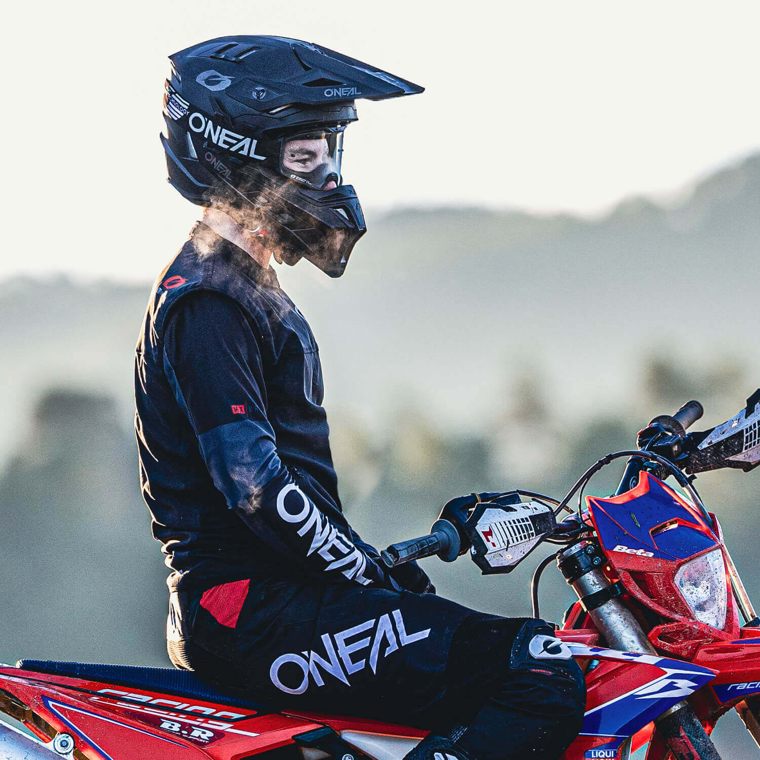 Eine Person mit schwarzem O'Neal-Helm und Rennausrüstung sitzt auf einem rot-blauen Dirtbike und blickt zur Seite. Sie scheint sich im Freien zu befinden, mit einem unscharfen natürlichen Hintergrund, möglicherweise Bäumen und Hügeln.