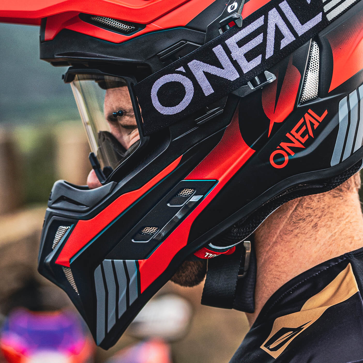Nahaufnahme einer Person, die einen leuchtend roten und schwarzen Motocrosshelm trägt, auf dem deutlich der Markenname „Oneal“ zu sehen ist. Der Helm hat ein schlankes, aerodynamisches Design mit detaillierten Markierungen und einem klaren Visier. Der Hintergrund ist leicht verschwommen.
