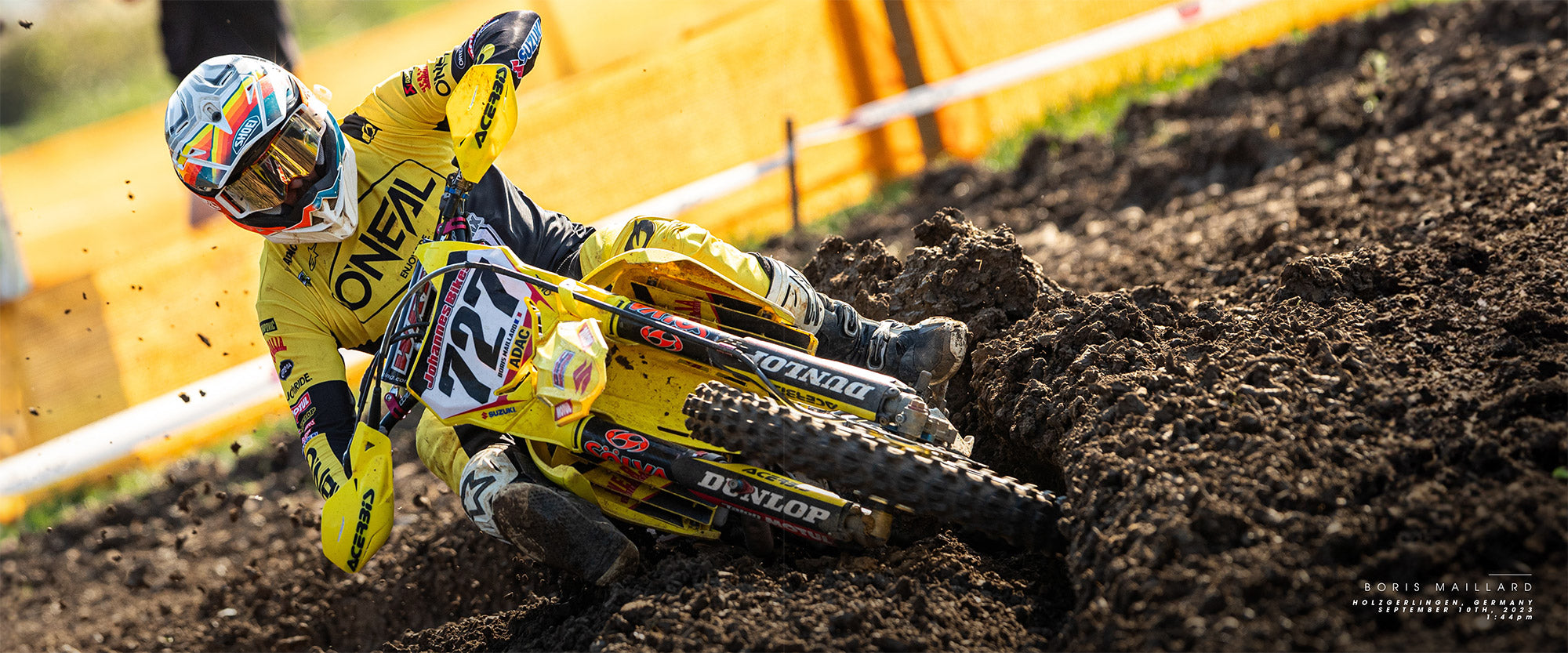 Ein Motocrossfahrer in einem gelben O'Neal-Outfit und Helm manövriert ein Dirtbike präzise über eine schlammige Strecke. Der Hintergrund ist verschwommen, was Bewegung und Geschwindigkeit andeutet. Der Fahrer konzentriert sich darauf, in dem unwegsamen Gelände Gleichgewicht und Kontrolle zu halten.