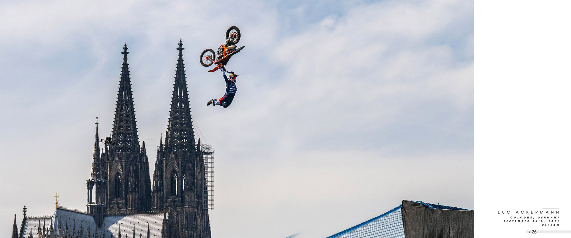 Vor der Kulisse der beiden Türme des Kölner Doms vollführt ein Motocross-Fahrer einen Stunt in der Luft. Der Fahrer lässt kopfüber sein Bike los und zeigt einen beeindruckenden Trick. Der Himmel ist teilweise bewölkt, was der Action-Aufnahme einen dramatischen Effekt verleiht.