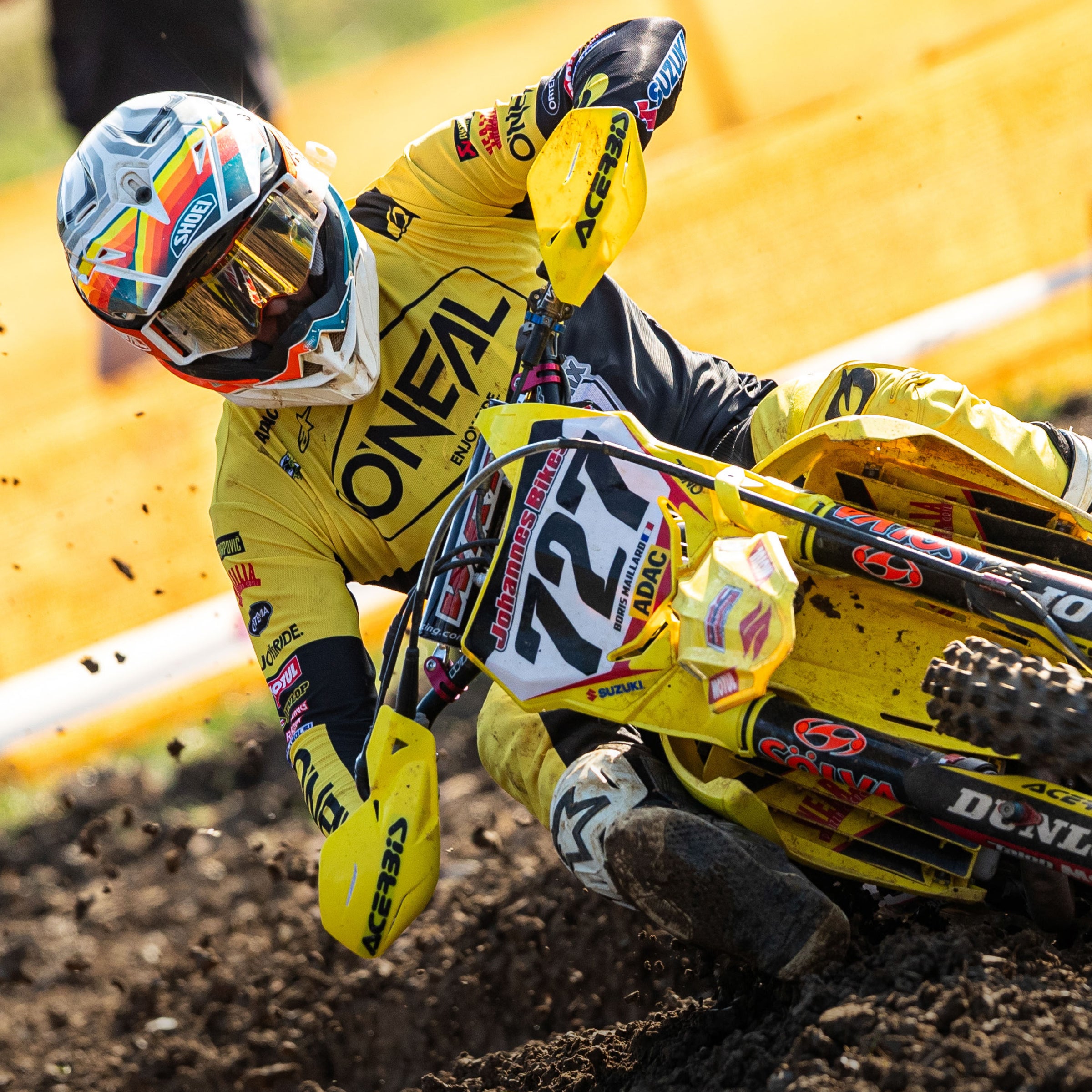 Ein Motocrossfahrer in gelbem Outfit und Helm mit buntem Visier mit der Nummer 127 legt sich auf einer unbefestigten Piste in eine scharfe Kurve, sodass Staub um das Motorrad herumwirbelt. Der Fahrer hält die Lenker fest und zeigt Konzentration und Kontrolle.