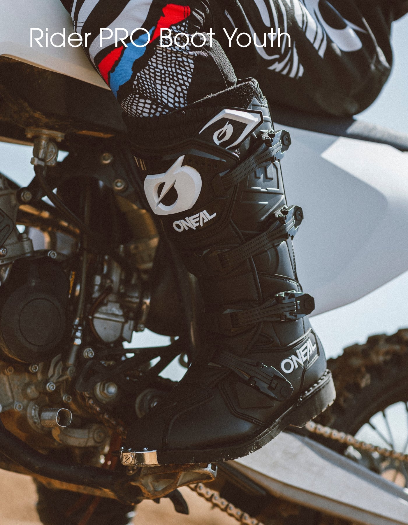 Nahaufnahme einer Person in schwarzen O'NEAL Rider PRO Motocross-Stiefeln, die ihren Fuß auf die Fußraste eines Geländemotorrads stützt. Der Text auf dem Bild lautet "Rider PRO Boot Youth".