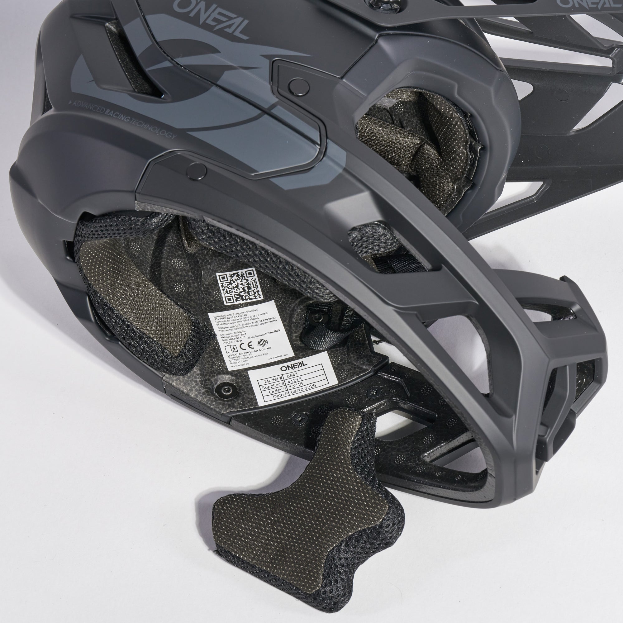 Nahaufnahme eines schwarz-grauen O'Neal-Fahrradhelms, die die Innenpolsterung, den Kinnschutz und ein QR-Code-Etikett im Inneren des Helms zeigt. Der Helm ruht auf einer weißen Fläche.