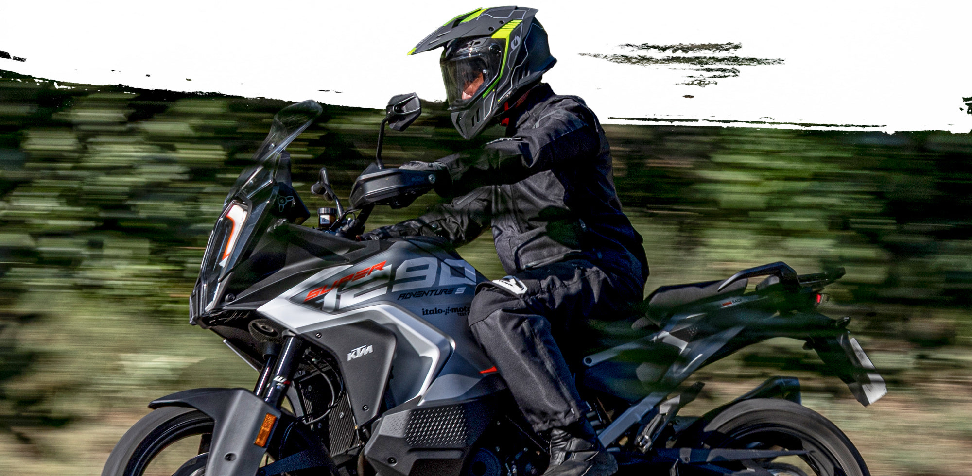 Eine Person in schwarzer Motorradkleidung und mit Helm fährt ein graues und schwarzes KTM 890 Adventure-Motorrad auf einer Straße mit Grünflächen im Hintergrund.