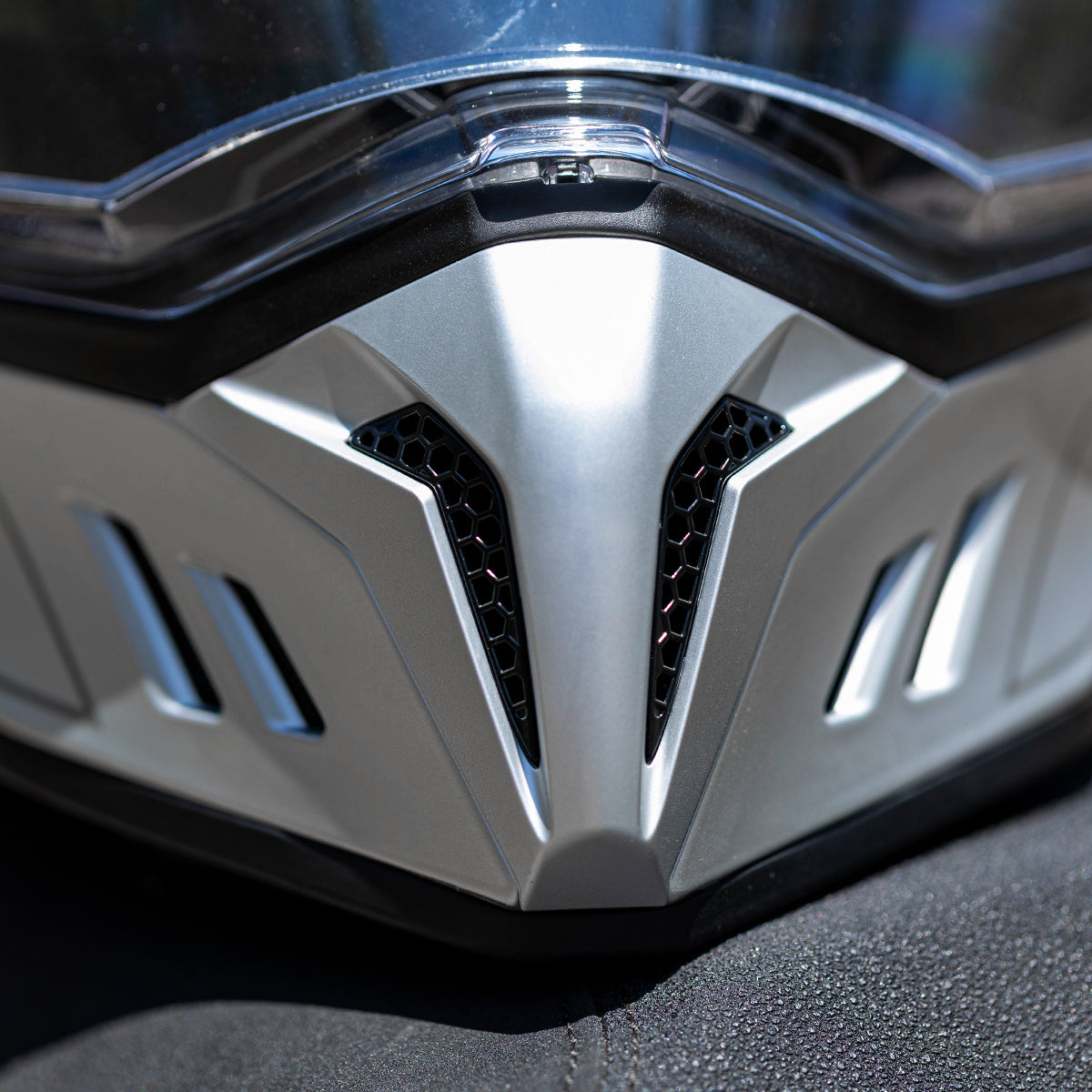 Nahaufnahme eines futuristischen, silbernen Motorradhelms mit Schwerpunkt auf dem kantigen Design der vorderen Belüftungsöffnungen mit schwarzem Netz unter einem klaren Visier.