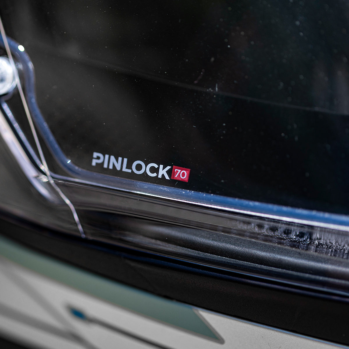 Nahaufnahme eines durchsichtigen Helmvisiers mit aufgedrucktem "PINLOCK 70"-Logo, das einen Teil der Außenschale des Helms und eine Reflexion auf der Visieroberfläche zeigt.