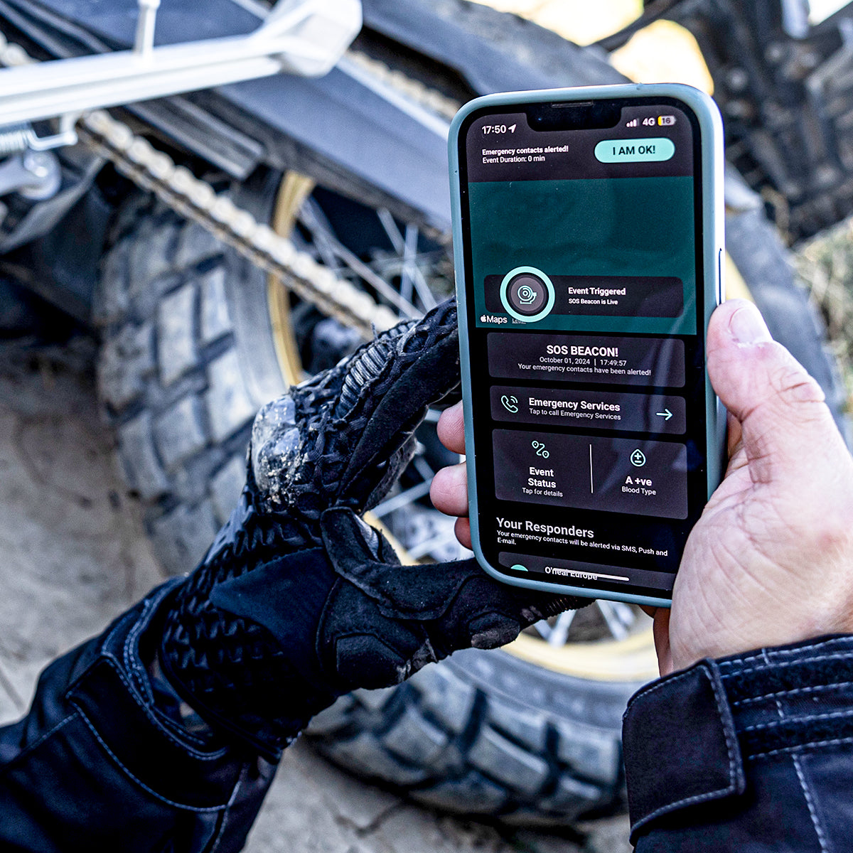 Eine Person mit schwarzen Handschuhen hält ein Smartphone in der Hand, auf dem eine SOS-Notfall-App angezeigt wird, während im Hintergrund ein Motorrad und sandiger Boden zu sehen sind.