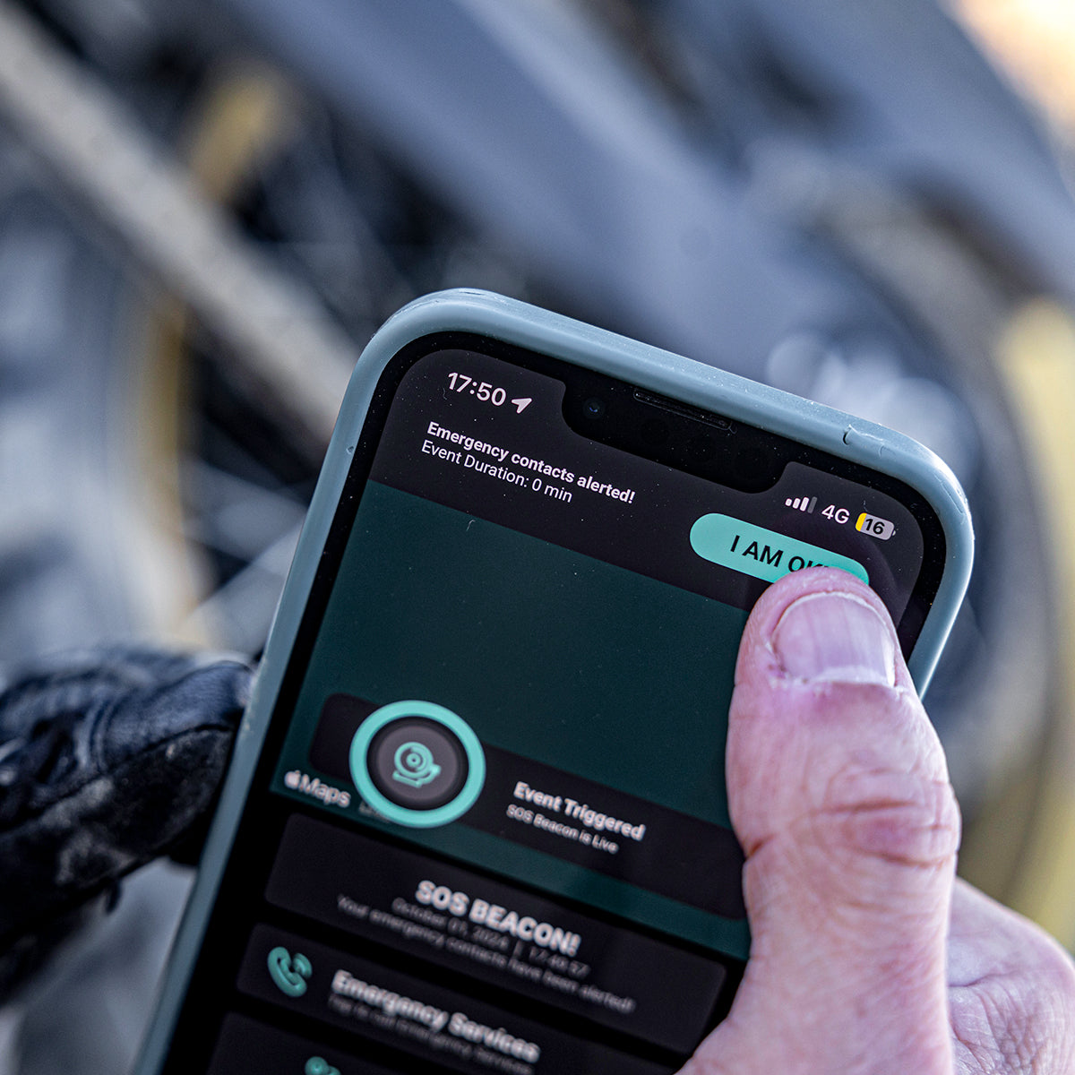 Eine behandschuhte Hand hält ein Smartphone mit einer SOS-Alarm-App, auf deren Bildschirm "Notfallkontakte alarmiert" und "Ereignis ausgelöst" angezeigt wird. Das Telefon befindet sich in der Nähe eines Fahrradrads im unscharfen Hintergrund.