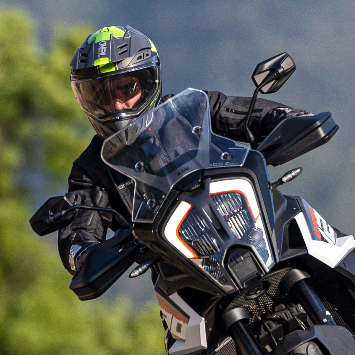 Eine Person, die eine schwarze Jacke und einen Helm mit grünen Akzenten trägt, fährt ein schwarz-weißes Motorrad und biegt auf einer Straße mit unscharfem Naturhintergrund in eine Kurve ein.