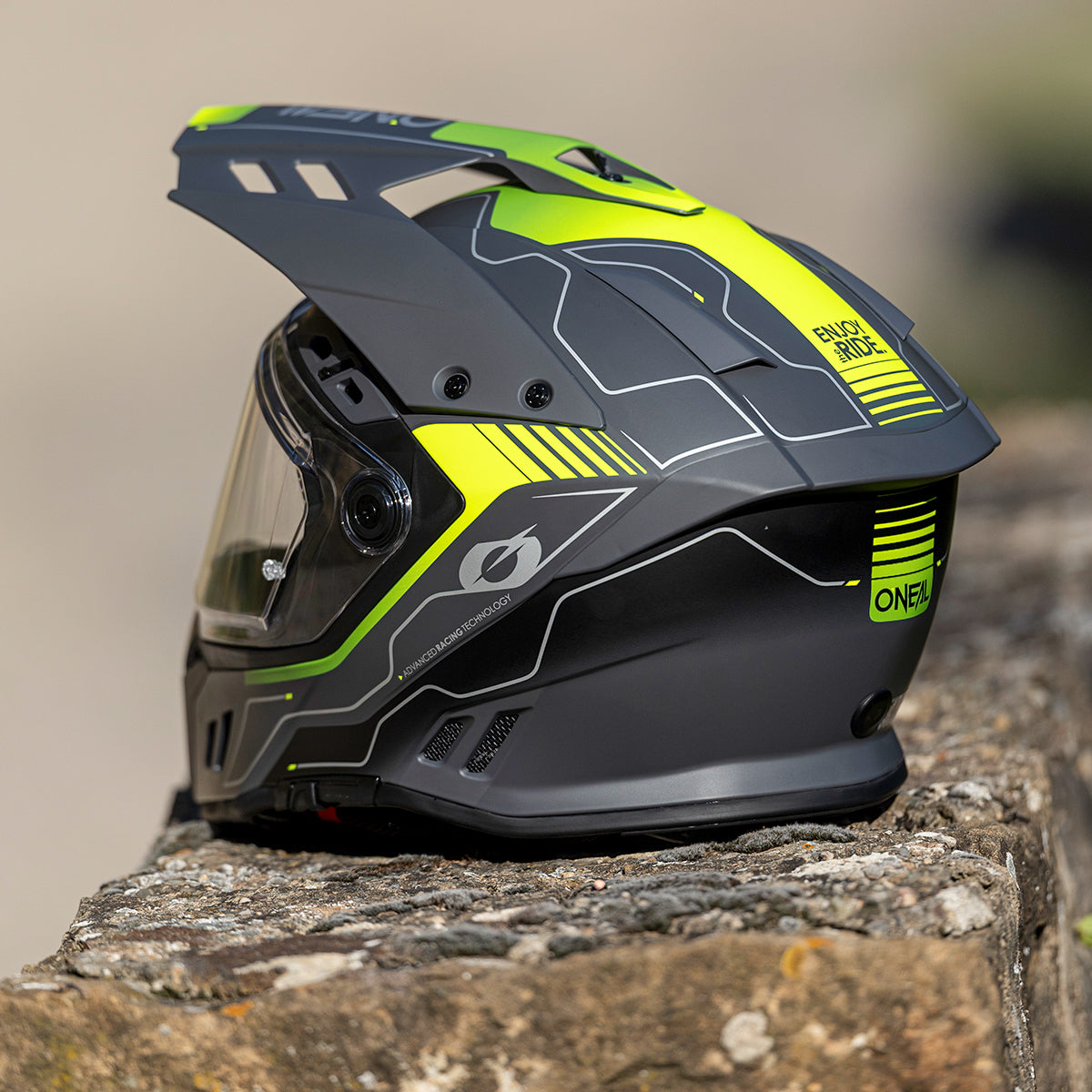 Ein schwarzer und neongelber Motorradhelm mit geometrischen Mustern ruht im Freien auf einer Steinoberfläche, von der Seite betrachtet. Der Helm verfügt über ein klares Visier und Belüftungsdetails.