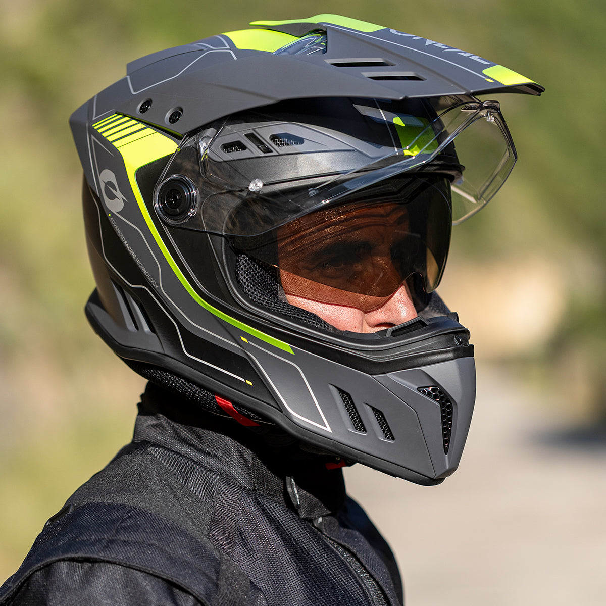 Eine Person, die einen schwarzen und neongelben Motorradhelm mit durchsichtigem Visier und schwarzer Fahrkleidung trägt, wird im Profil vor einem unscharfen Hintergrund im Freien gezeigt.