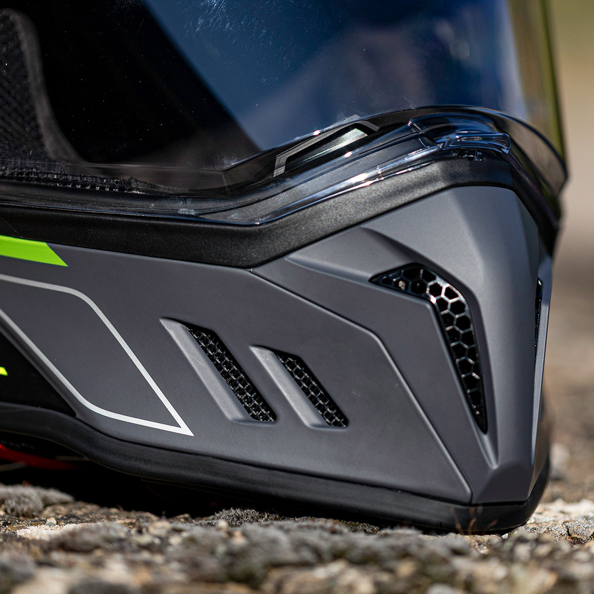 Nahaufnahme des Kinnbereichs eines schwarz-grauen Motorradhelms mit Belüftungsöffnungen, klarem Visier und ein paar neongelben Details, der auf einer felsigen Oberfläche im Freien liegt.