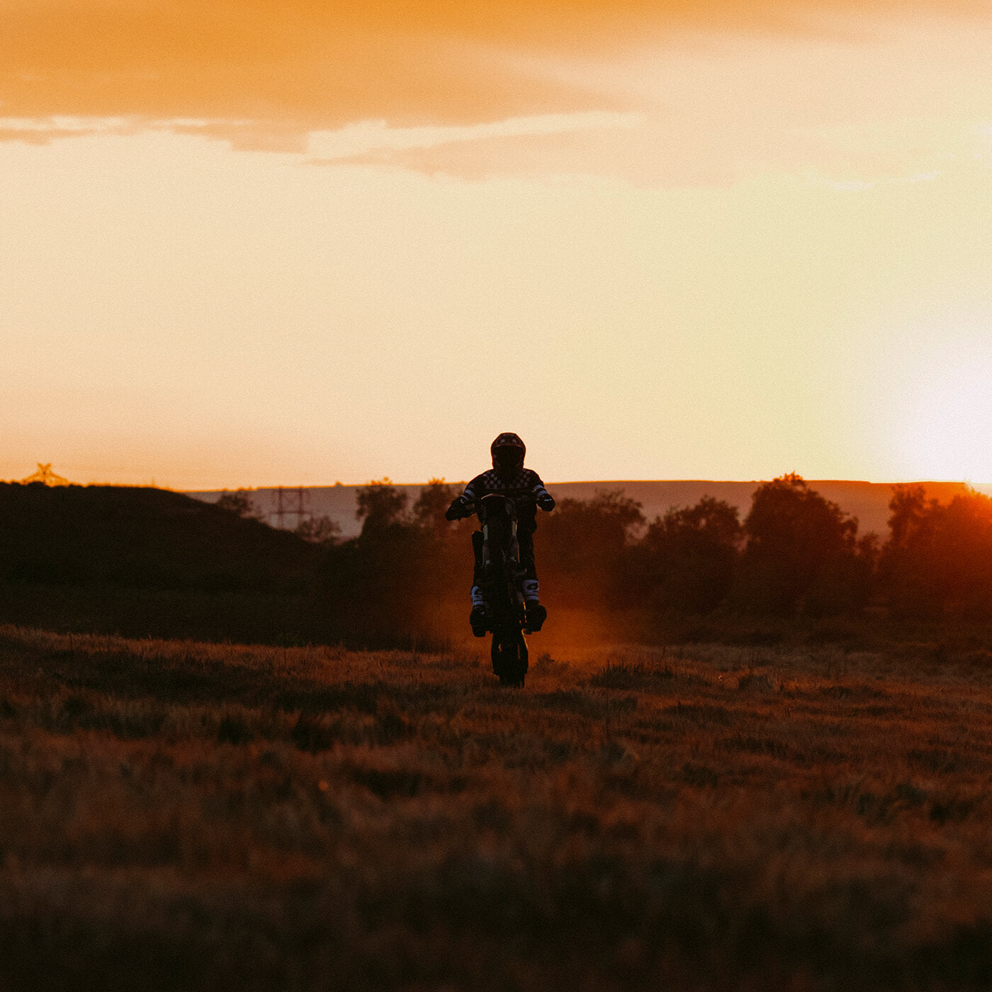 Ein Motorradfahrer macht einen Wheelie auf einem Dirtbike, während er bei Sonnenuntergang durch ein offenes Feld fährt. Der Himmel ist orange und golden, die Sonne ist teilweise am Horizont sichtbar und wirft einen warmen Schein über die Landschaft. Im Hintergrund sind Bäume und entfernte Hügel als Silhouetten zu erkennen.