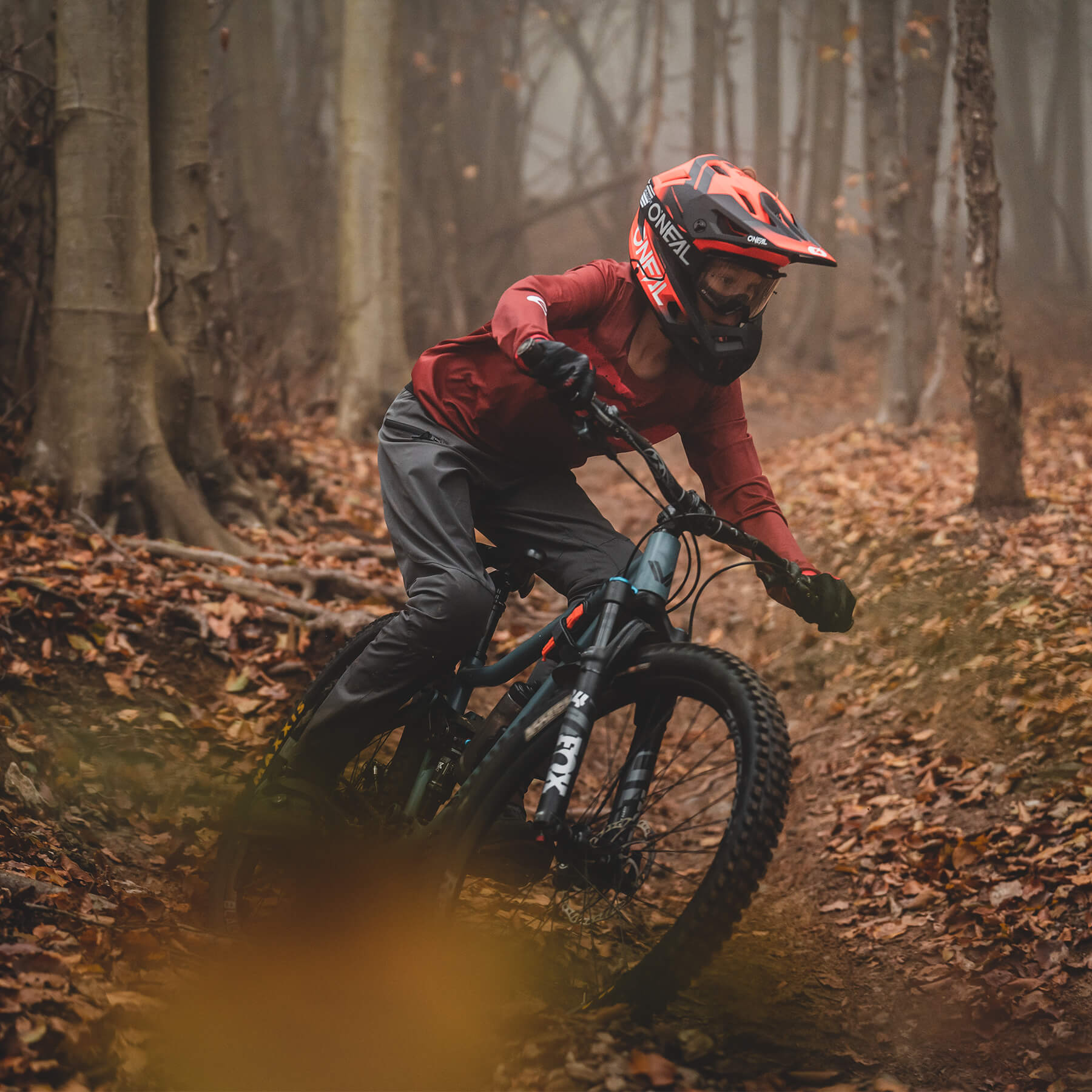 Eine Person mit rotem Helm, rotem Langarmshirt und schwarzer Hose fährt mit einem Mountainbike durch einen Waldweg, der mit Laub bedeckt ist. Die Umgebung ist neblig und der Weg ist schmal und auf beiden Seiten von Bäumen umgeben.
