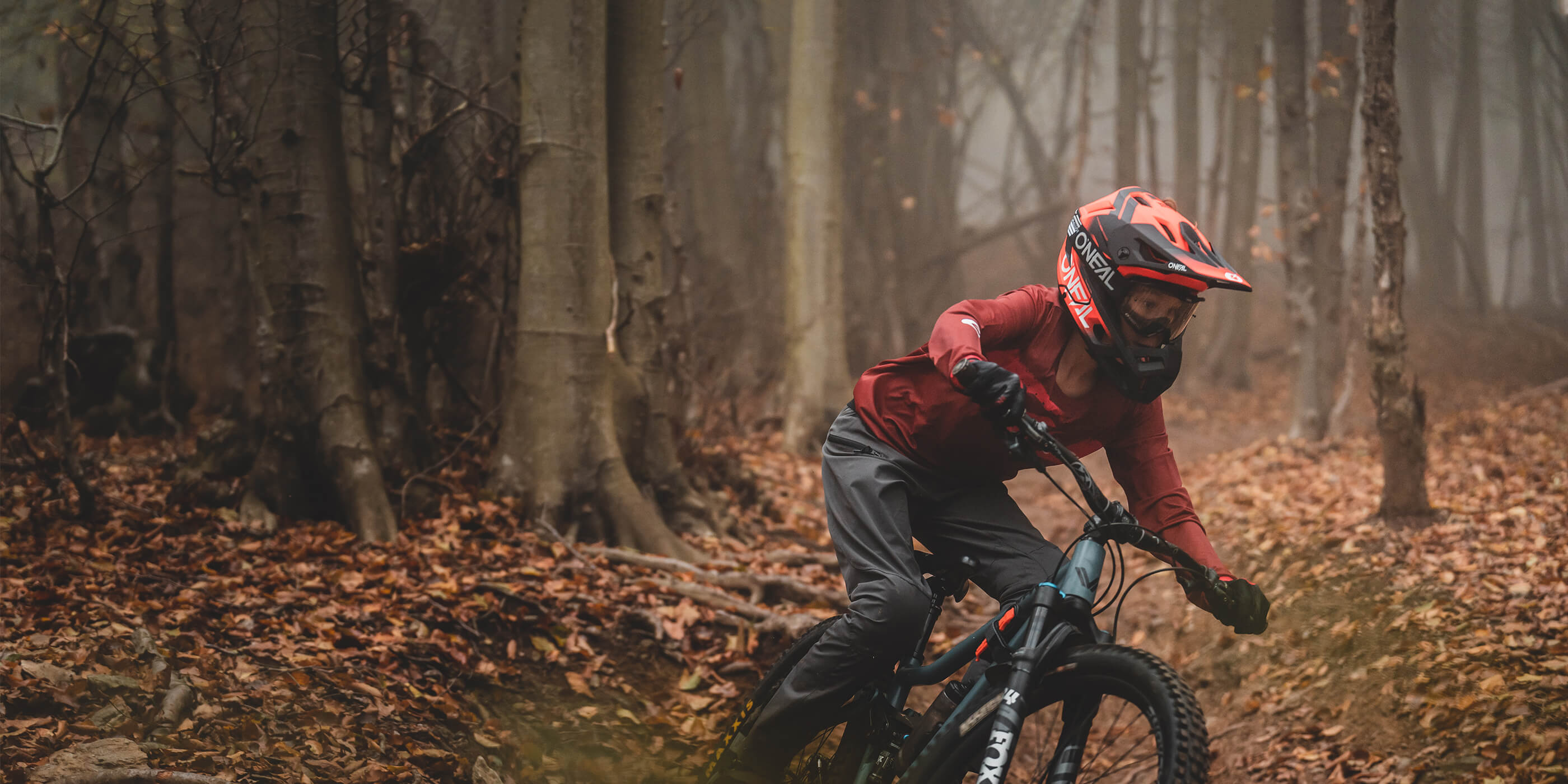Eine Person mit rotem Helm und Schutzausrüstung fährt mit einem Mountainbike durch einen dichten, nebligen Wald, dessen Boden mit Laub bedeckt ist. Sie navigiert einen Pfad entlang, lehnt sich in eine Kurve und ist von hohen Bäumen mit kahlen Ästen umgeben.
