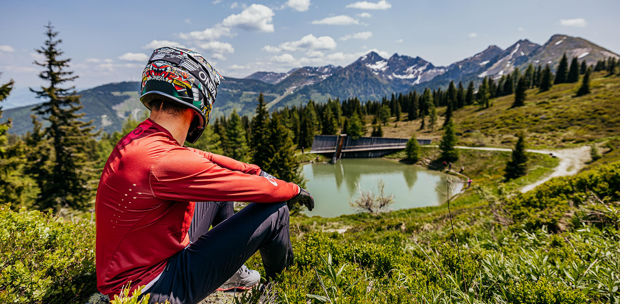 Eine Person mit einem bunten Helm und einem roten Langarmshirt sitzt auf einem grasbewachsenen Hügel und blickt auf eine ruhige Berglandschaft. Bäume umgeben einen kleinen See im Mittelgrund und im Hintergrund erheben sich schneebedeckte Berggipfel unter einem teilweise bewölkten Himmel.
