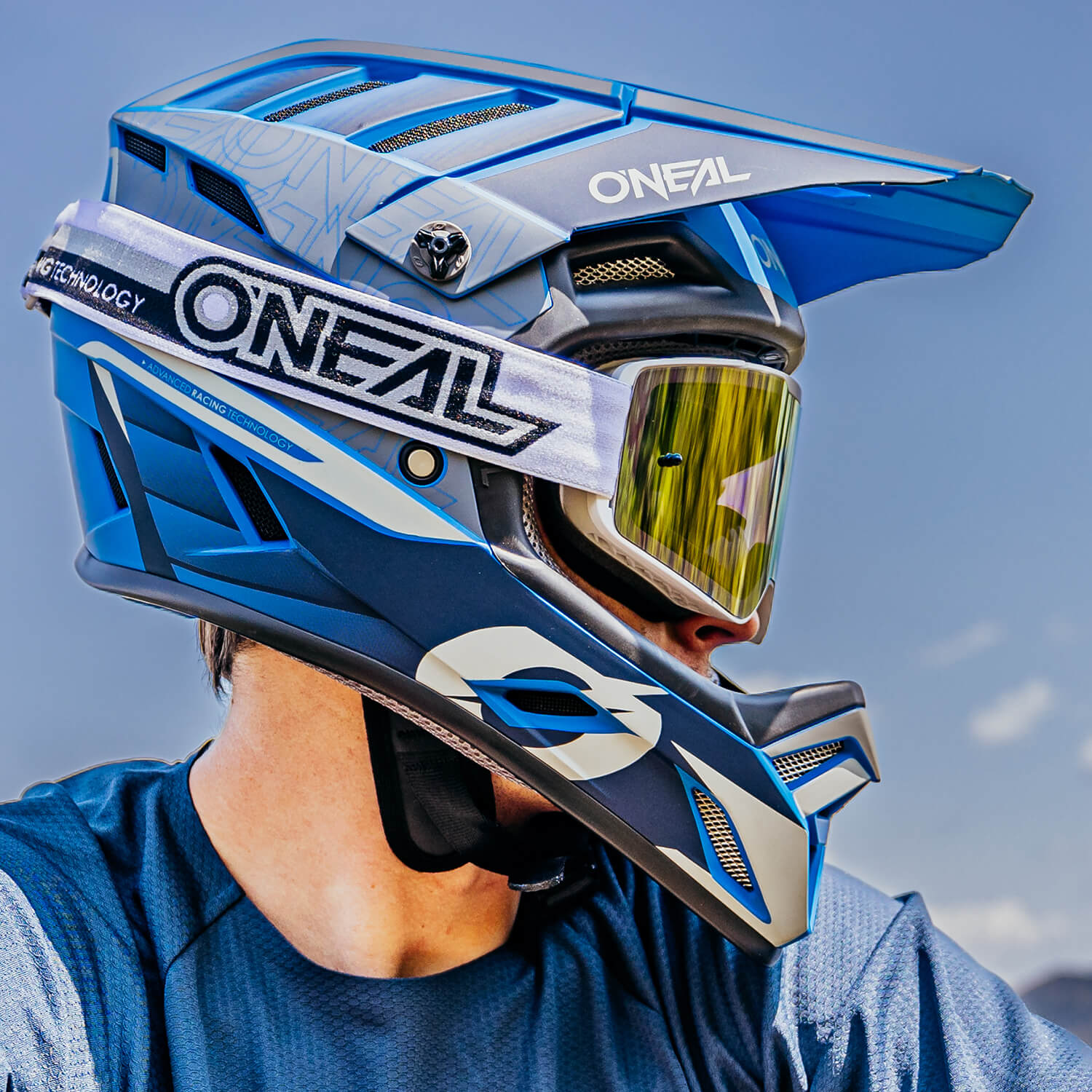 Nahaufnahme einer Person, die einen blau-weißen O'Neal-Motocrosshelm mit gelb getöntem Visier trägt und zur Seite blickt. Der Helm weist komplizierte Muster auf und das O'Neal-Logo ist deutlich zu sehen. Die Person trägt außerdem ein passendes blau-weißes Motocrosstrikot.
