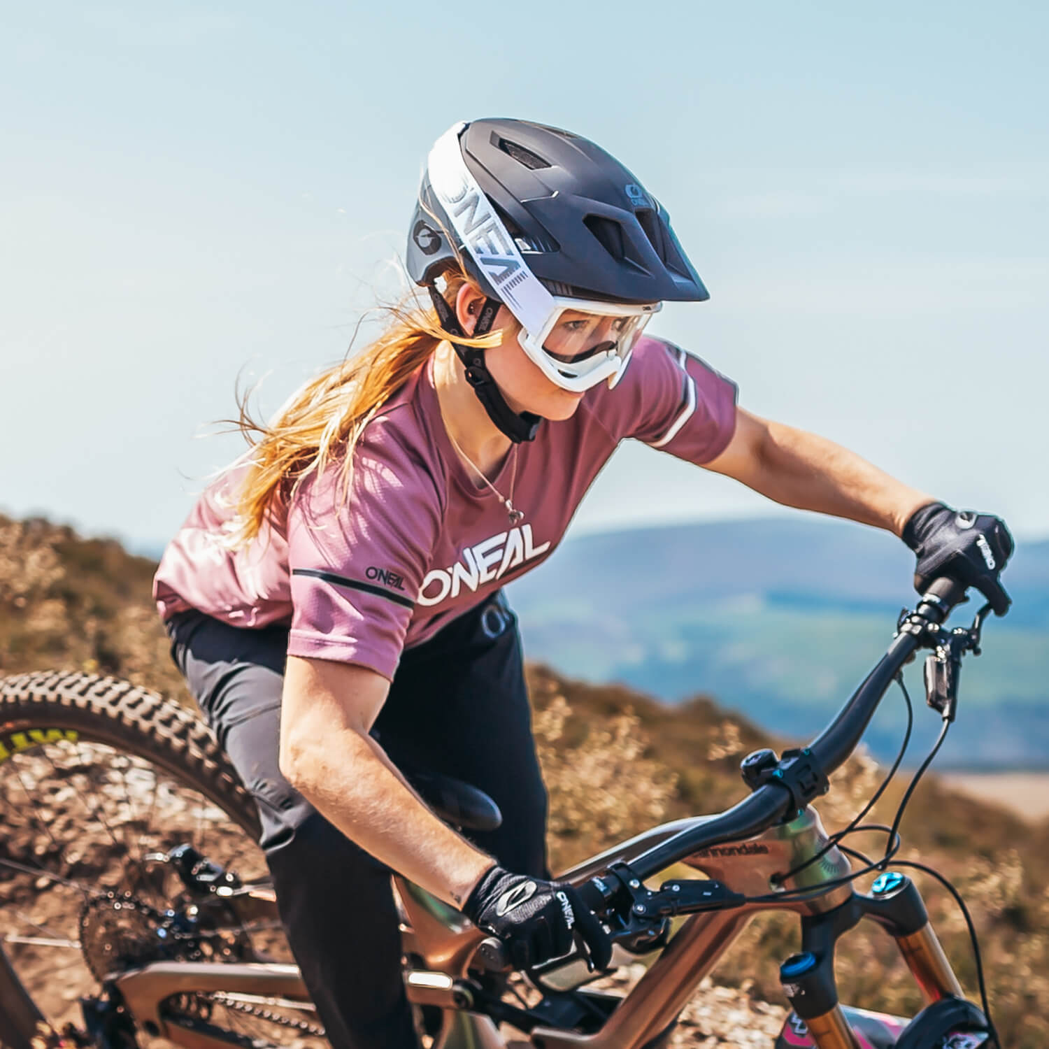 Eine Person mit Helm, Schutzbrille und rosa Radtrikot fährt mit einem Mountainbike auf einem Feldweg. Im Hintergrund ist eine malerische Landschaft mit Hügeln und klarem Himmel zu sehen. Die Person lehnt sich nach vorne und konzentriert sich darauf, den Weg zu finden.