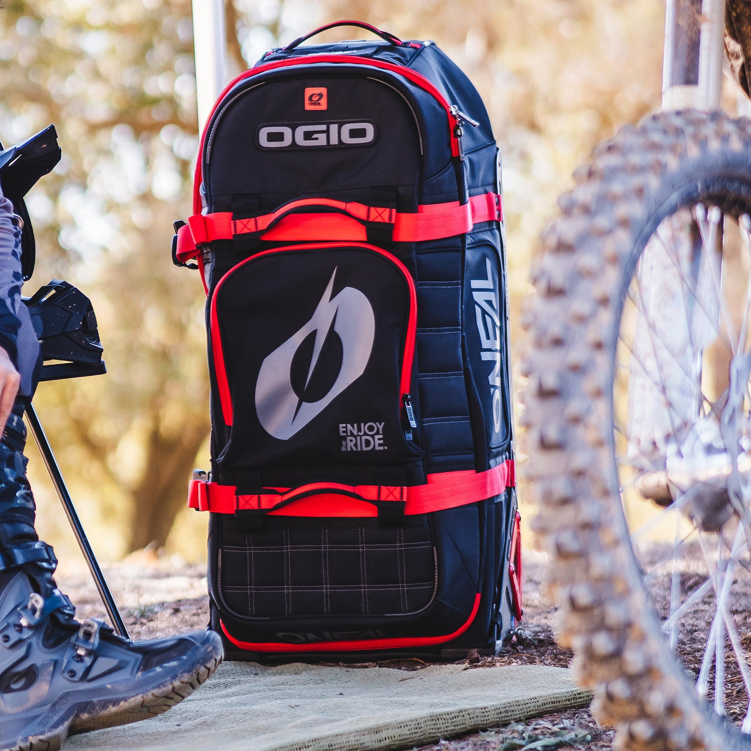 Eine schwarz-rote OGIO-Sporttasche mit dem Logo und den Worten „Enjoy the Ride“. Ein Paar Motocross-Stiefel und ein Teil einer Person sind zu sehen. Im Vordergrund ist ein Motocross-Rad zu sehen, im Hintergrund sind verschwommene Bäume zu sehen.