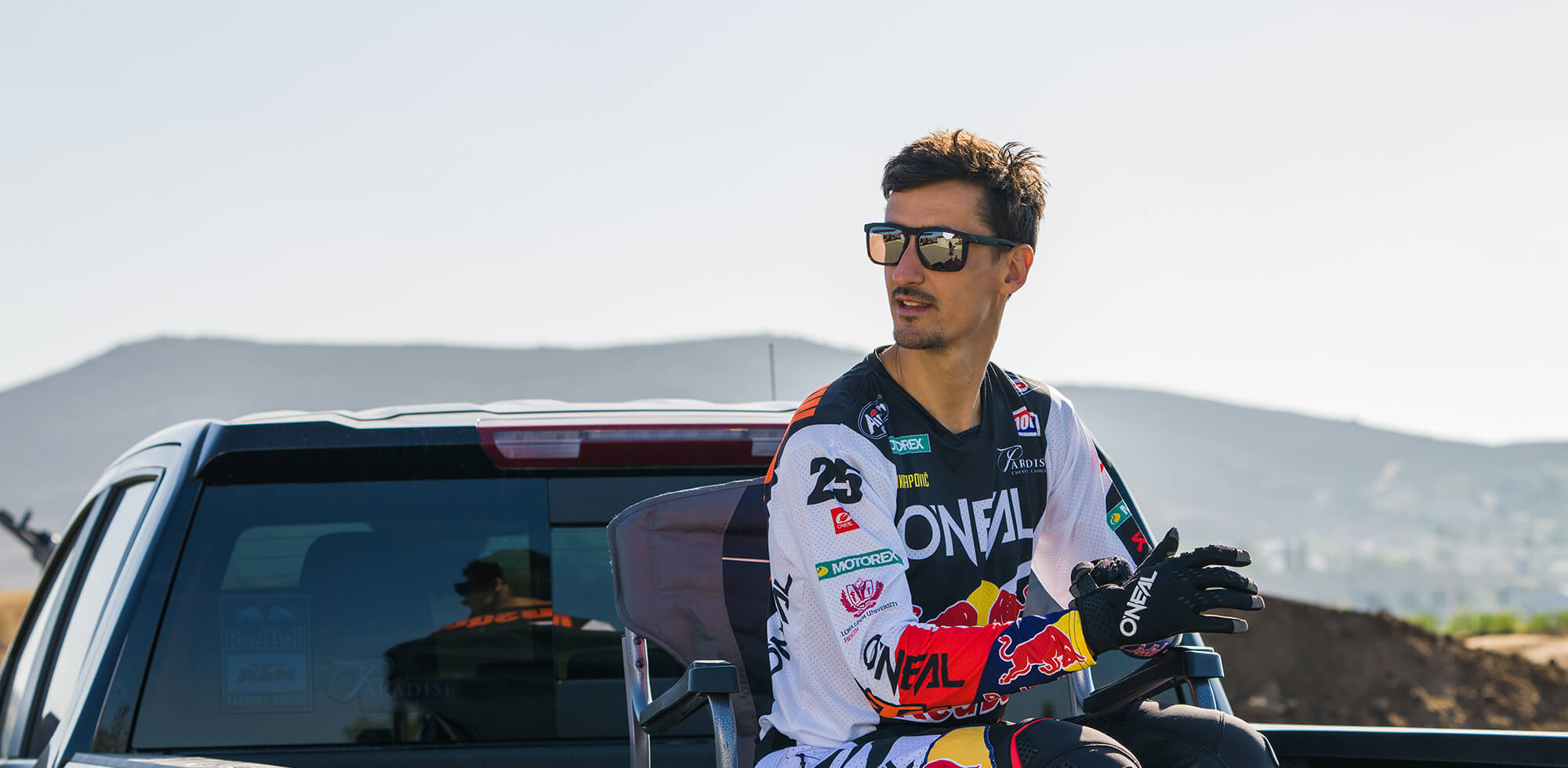 Eine Person in Motocross-Outfit, Sonnenbrille und Handschuhen sitzt auf der Ladefläche eines Pickups. Im Hintergrund ist ein sonniger Tag mit sanften Hügeln und klarem Himmel zu sehen. Die Person blickt in die Ferne, bereitet sich möglicherweise auf eine Fahrt vor oder macht eine Pause.