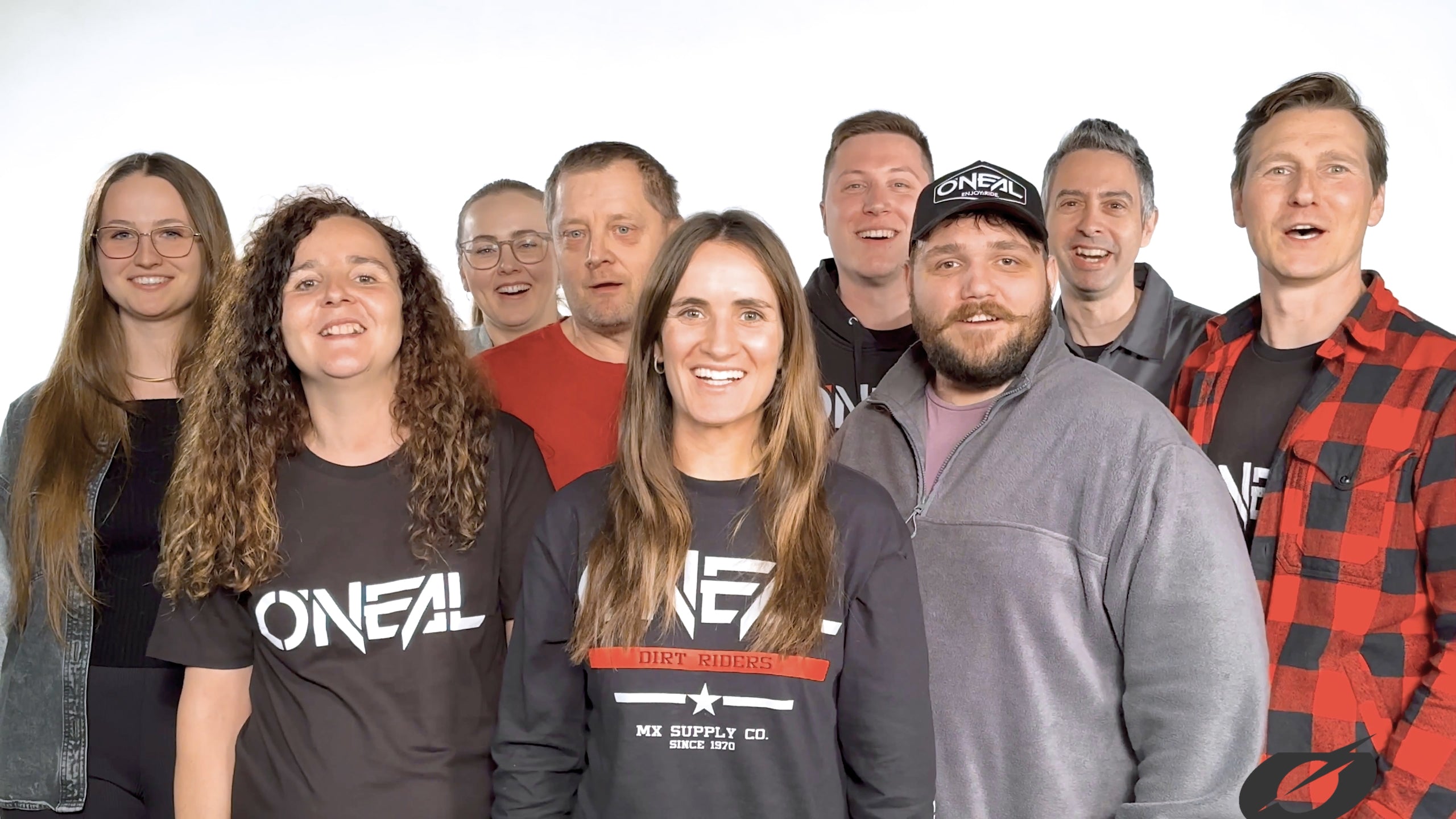 Eine Gruppe von neun Personen, Männern und Frauen, steht lächelnd zusammen. Einige tragen T-Shirts und Hüte mit dem Logo von O’Neal. Der Hintergrund ist weiß und hell und sorgt für eine fröhliche und freundliche Atmosphäre.