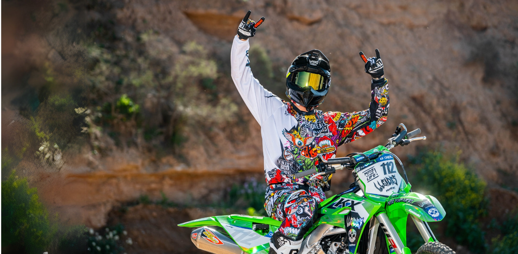 Ein Motocross-Fahrer in buntem Anzug und Helm sitzt auf einem grünen Motorrad mit der Nummer 112 und hebt vor einem felsigen Hintergrund im Freien beide Arme und Hände in der Geste „Rock on“.