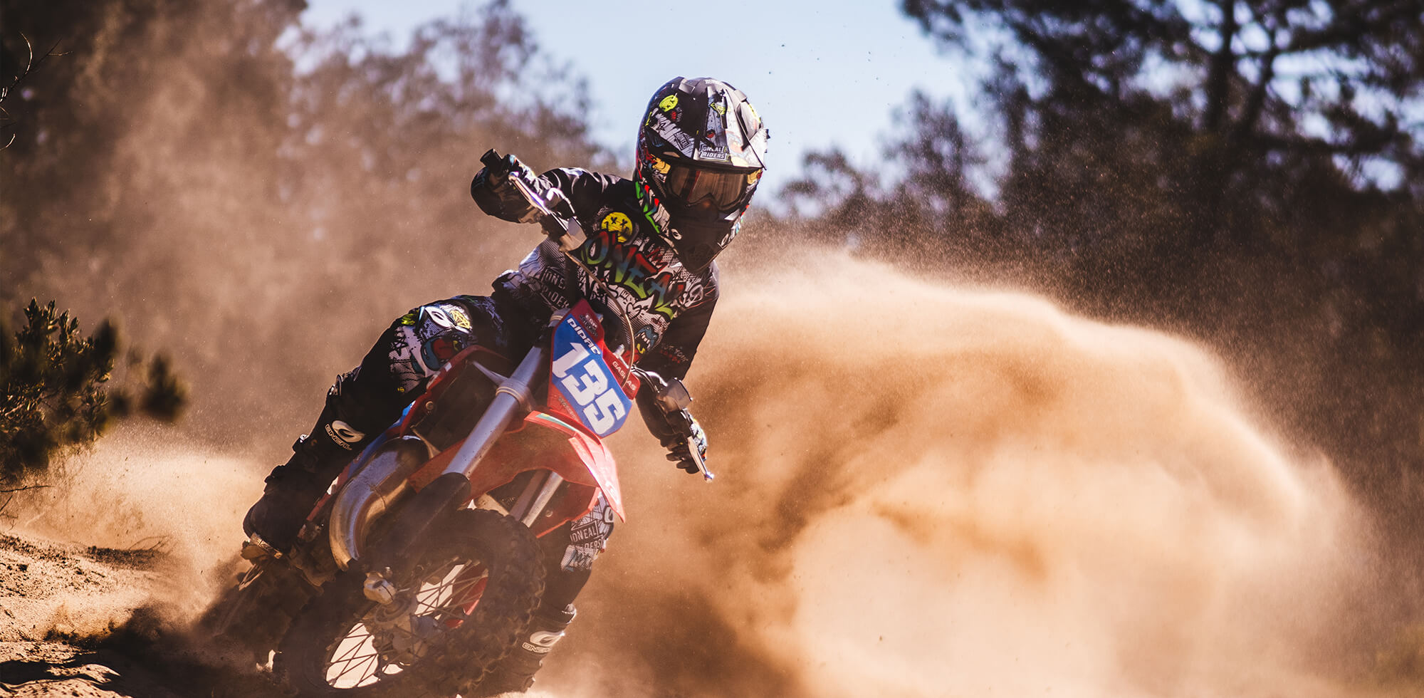 Ein Motocrossfahrer auf dem Motorrad Nummer 136 fährt eine scharfe Kurve auf einer unbefestigten Strecke und wirbelt dabei eine Staubwolke auf. Der Fahrer trägt einen Helm und eine komplette Montur, die mit verschiedenen bunten Aufklebern verziert ist. Im Hintergrund sind Bäume und ein klarer blauer Himmel zu sehen.