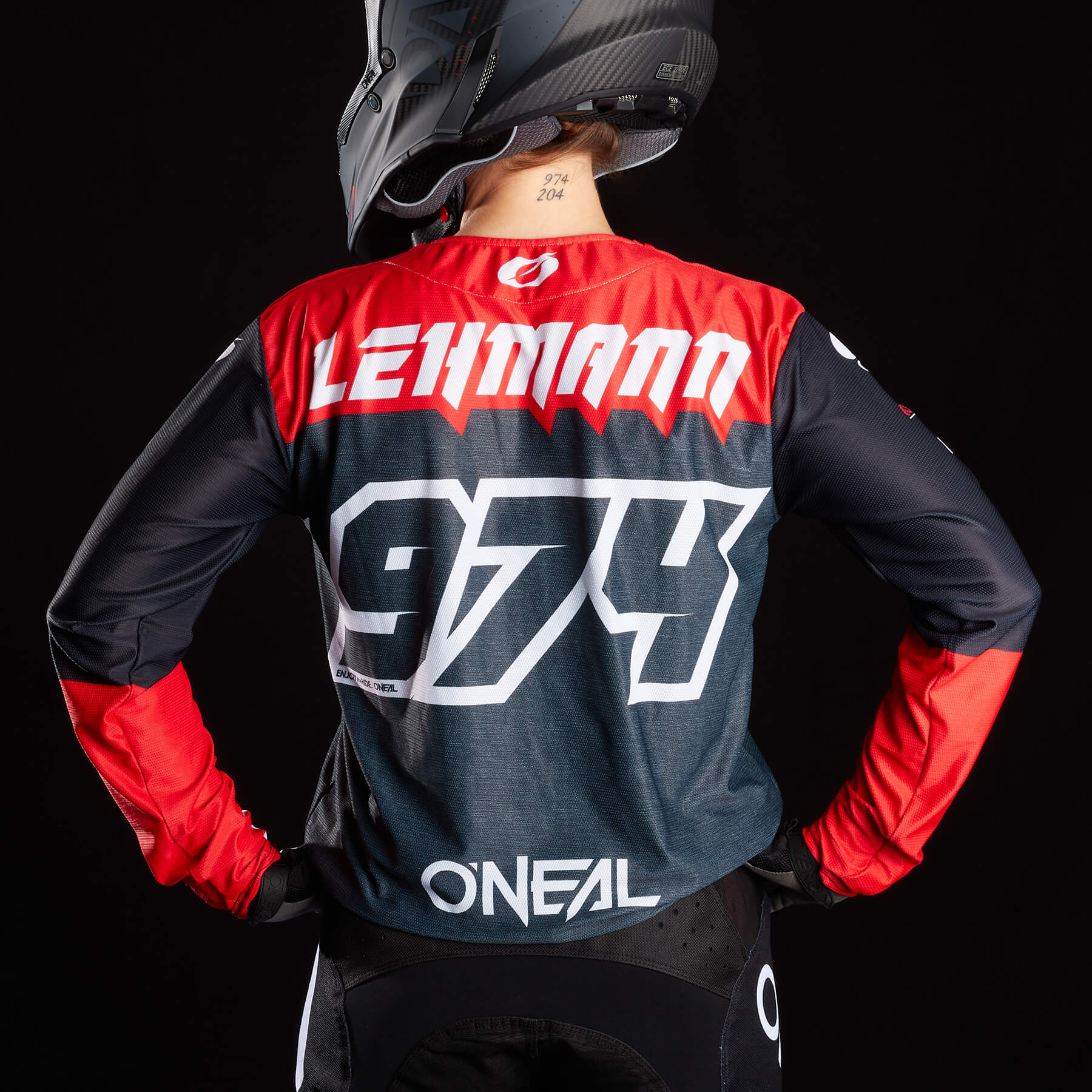 Eine Person in einem schwarz-roten Motocross-Trikot mit „LEHMANN“ und der Nummer „974“ in fetten weißen Buchstaben auf dem Rücken steht mit den Händen in den Hüften. Sie trägt außerdem Schutzkleidung, darunter einen Helm, vor einem schwarzen Hintergrund.