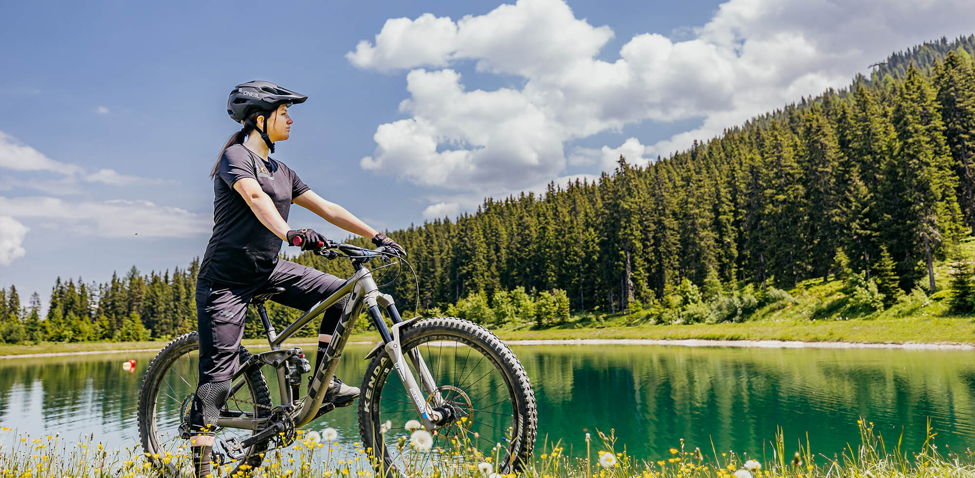 Eine Person mit Helm und Radsportausrüstung steht mit einem Mountainbike neben einem ruhigen See. Der See ist von üppigem Grün und hohen Kiefern umgeben, der Himmel ist teilweise bewölkt und das Sonnenlicht scheint hell.