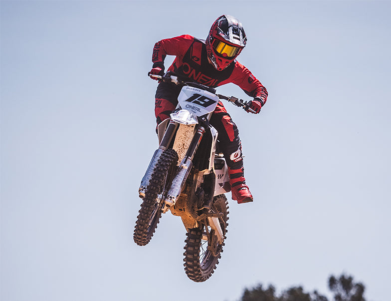 Ein Motocrossfahrer in voller roter Montur, einschließlich Helm und Schutzbrille, holt vor einem klaren blauen Himmel mitten im Sprung auf einem Dirtbike Luft. Das Motorrad und die Montur sind mit Markenzeichen versehen und die Haltung des Fahrers lässt auf Geschwindigkeit und Können schließen. Im Hintergrund sind Bäume zu sehen.
