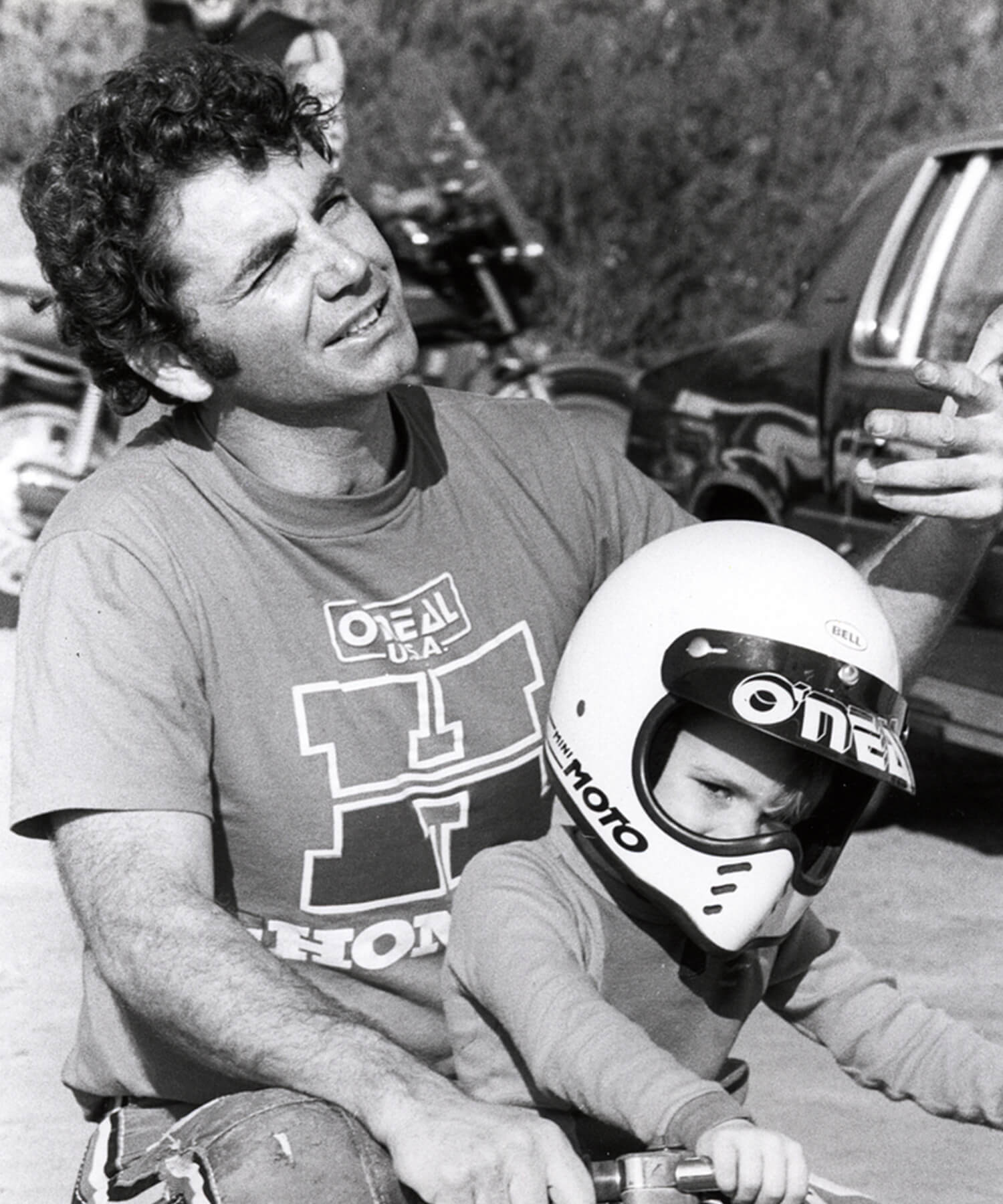 Ein Mann mit lockigem Haar und einem Hemd mit den Logos von O'Neal und Honda sitzt im Freien neben einem Kind, das einen Motorradhelm mit der Aufschrift „MOTO“ trägt. Das Kind scheint auf einem kleinen Motorrad zu sitzen. Im Hintergrund sind Fahrzeuge und Bäume zu sehen.