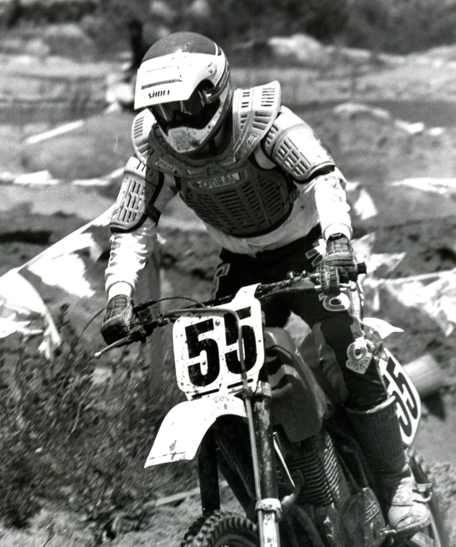 Ein Motocrossfahrer mit Helm, Schutzausrüstung und Trikot fährt ein Dirtbike mit der Nummer 55 auf dem vorderen Kennzeichen. Der Fahrer fährt auf einer holprigen Outdoor-Strecke mit markierten Begrenzungen. Das Bild ist schwarzweiß.