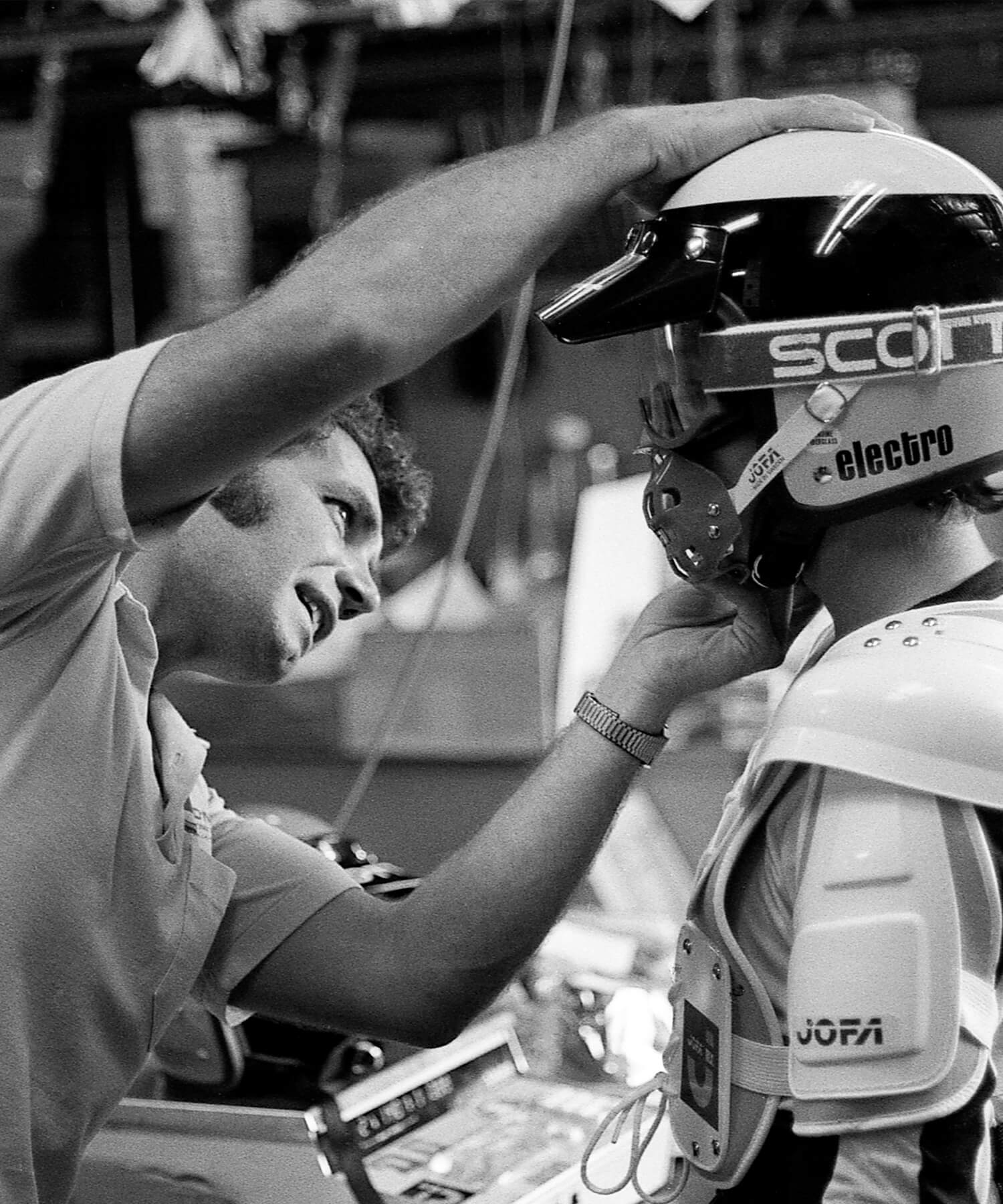 Ein Mann hilft einem Hockeyspieler, seinen Helm zurechtzurücken. Die Szene scheint eine Umkleidekabine oder ein Ausrüstungsbereich zu sein, im Hintergrund sind verschiedene Gegenstände zu sehen. Der Spieler trägt Schutzausrüstung, darunter Schulterpolster und einen Helm mit der Aufschrift „Scott“.