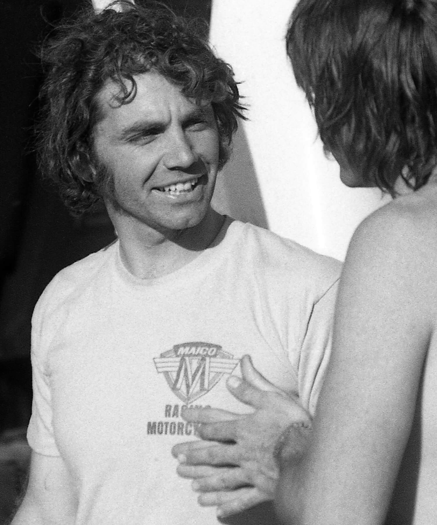 Auf einem Schwarzweißfoto sind zwei Männer im Gespräch zu sehen. Der Mann links hat welliges Haar und trägt ein helles Hemd mit dem Logo „Maico Racing Motorcycles“. Der Mann rechts ist oben ohne und hat längere Haare.