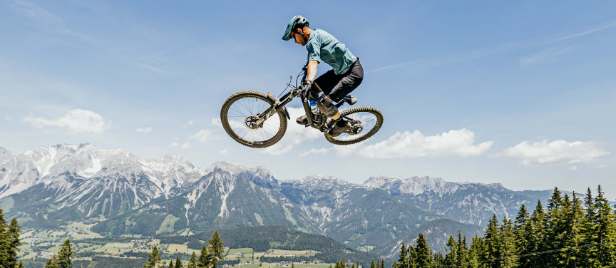 Eine Person mit Helm vollführt vor dem Hintergrund schroffer Berggipfel und eines klaren blauen Himmels einen beeindruckenden Sprung auf einem Mountainbike. Darunter sind bewaldete Hügel und Täler zu sehen. Die Person ist in der Luft und demonstriert in der malerischen Umgebung Geschick und Nervenkitzel.