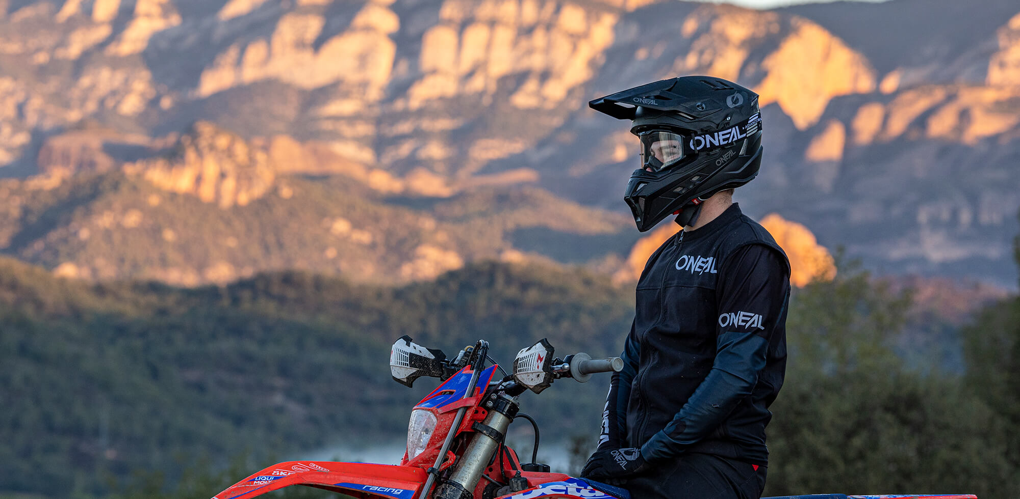 Eine Person mit schwarzem Helm und Motorradmontur sitzt auf einem roten Motorrad und blickt auf eine malerische Berglandschaft im Hintergrund. Der Himmel ist klar und die Sonne wirft einen warmen Schein auf das zerklüftete Gelände.
