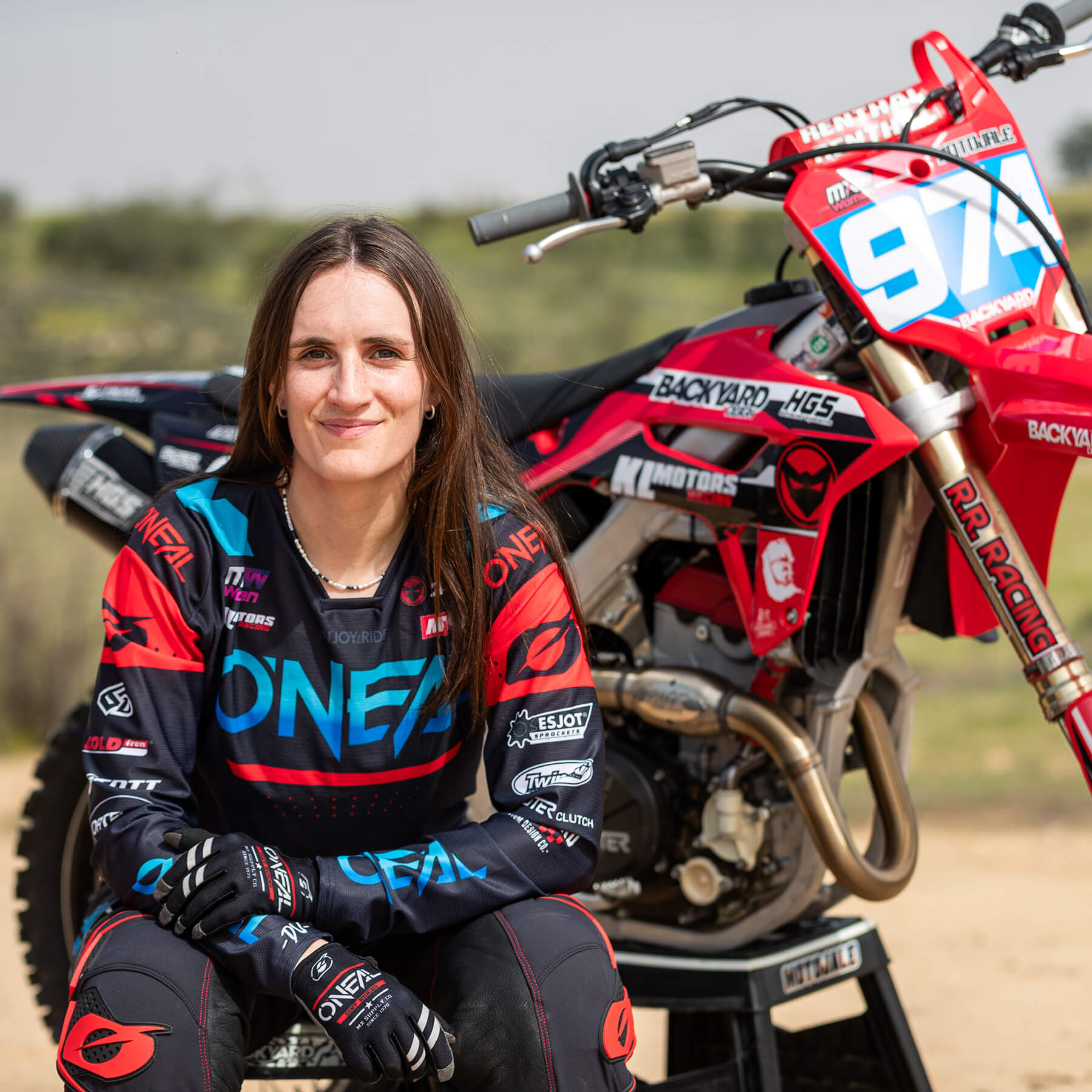 Eine Person mit langen Haaren sitzt auf einem Dirtbike-Ständer. Sie trägt ein komplettes Motocross-Outfit und Handschuhe und lächelt in die Kamera. Hinter ihr steht ein leuchtend rotes Dirtbike mit der Nummer 972 an prominenter Stelle. Der Hintergrund ist eine natürliche Umgebung im Freien.