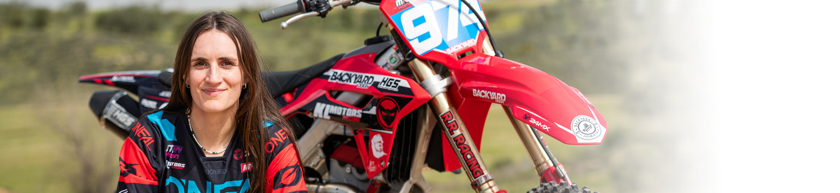 Eine Person mit langen dunklen Haaren und einem Motocross-Outfit sitzt lächelnd neben einem roten Dirtbike mit der Nummer 9 und verschiedenen Sponsorenlogos. Der Hintergrund ist eine Szene im Freien mit viel Grün, und die rechte Seite geht in einen weißen Farbverlauf über.