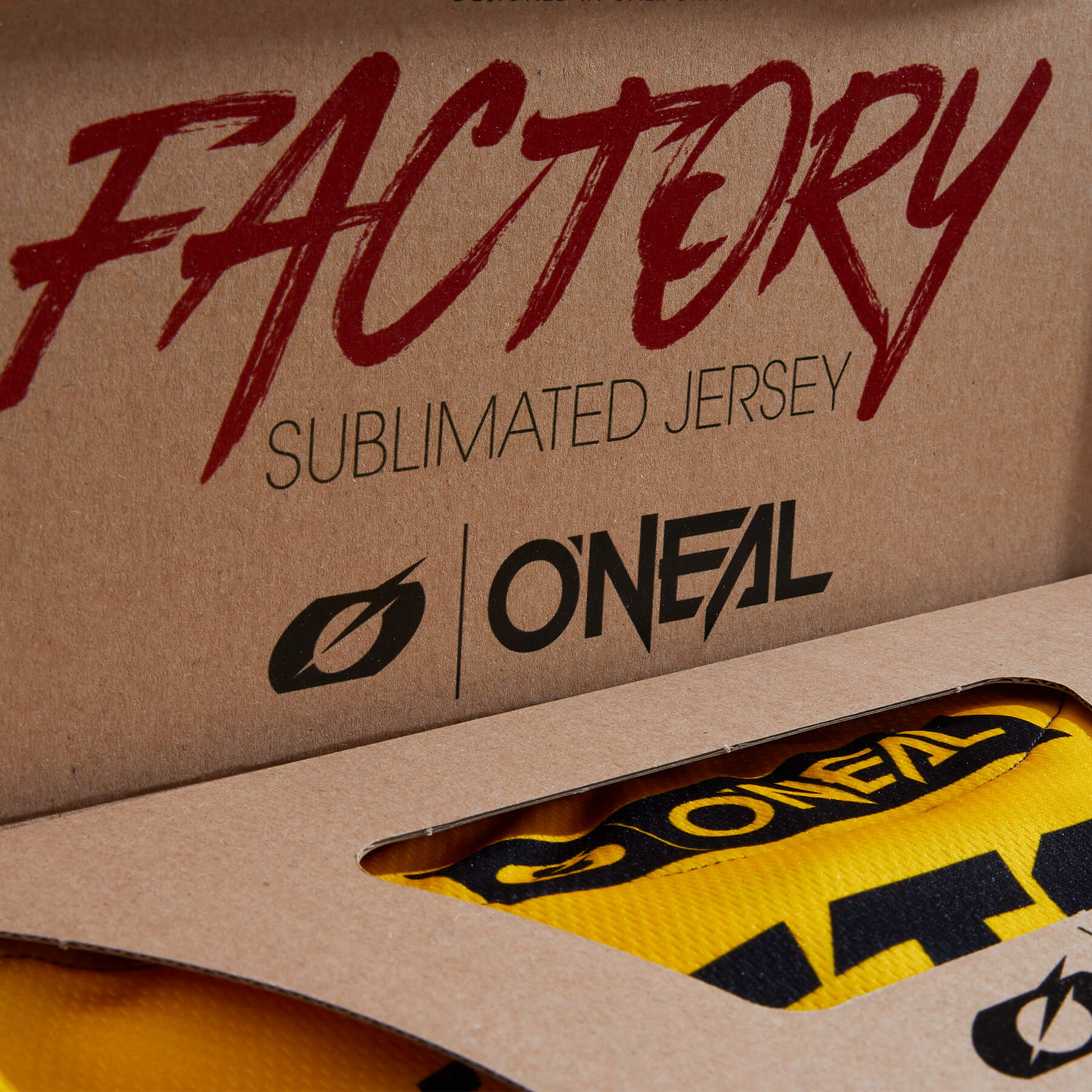 Eine Nahaufnahme eines Kartons mit den Worten „FACTORY Sublimated Jersey“ in roter Schrift und dem O'Neal-Logo darunter. Durch eine Öffnung im Karton ist ein Teil eines gelb-schwarzen Trikots sichtbar.