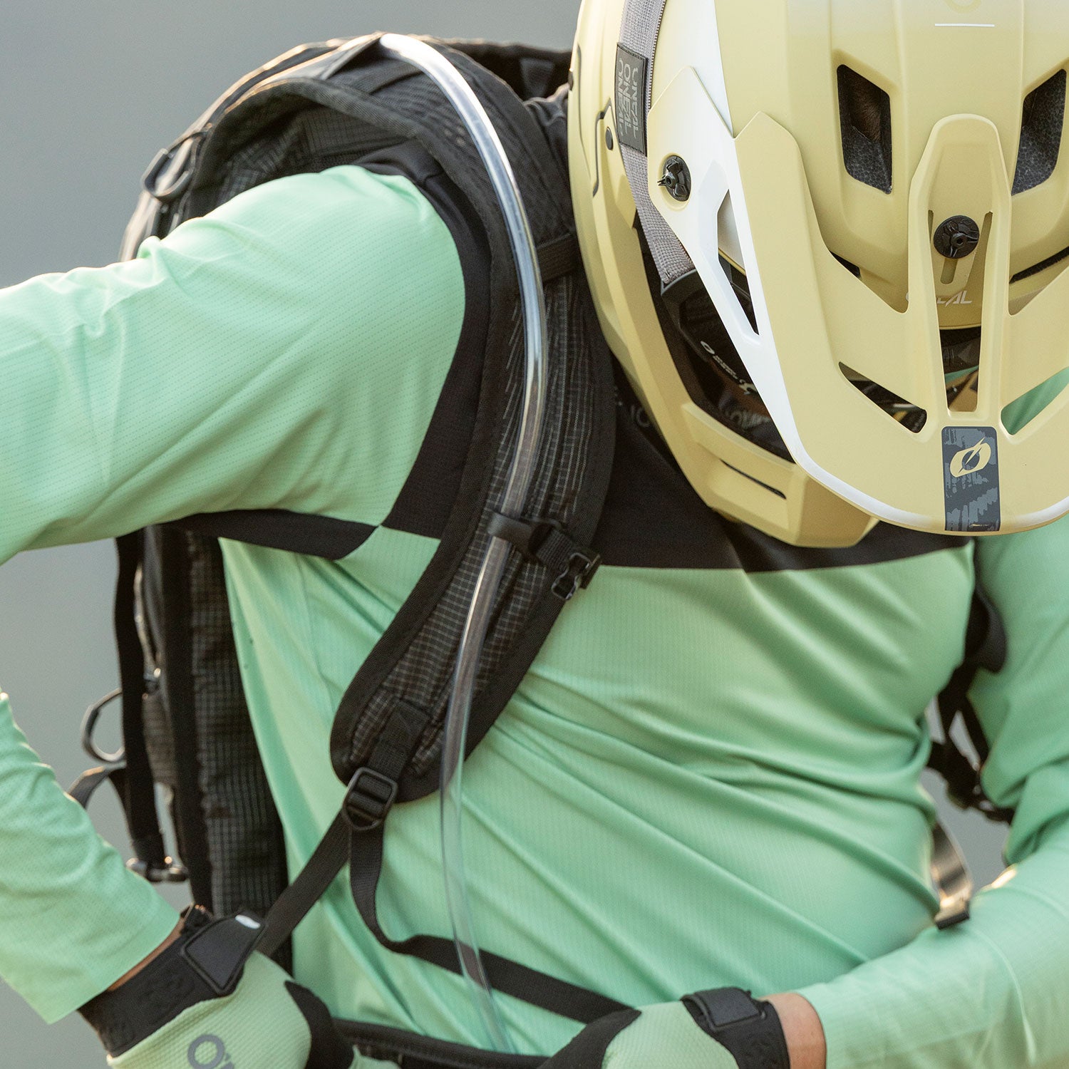 Eine Person, die ein hellgrünes langärmeliges Hemd, Handschuhe und einen beigen Fahrradhelm trägt, stellt die Gurte eines schwarzen Rucksacks mit einem Trinkschlauch ein. Das Bild ist so beschnitten, dass nur der Oberkörper zu sehen ist.