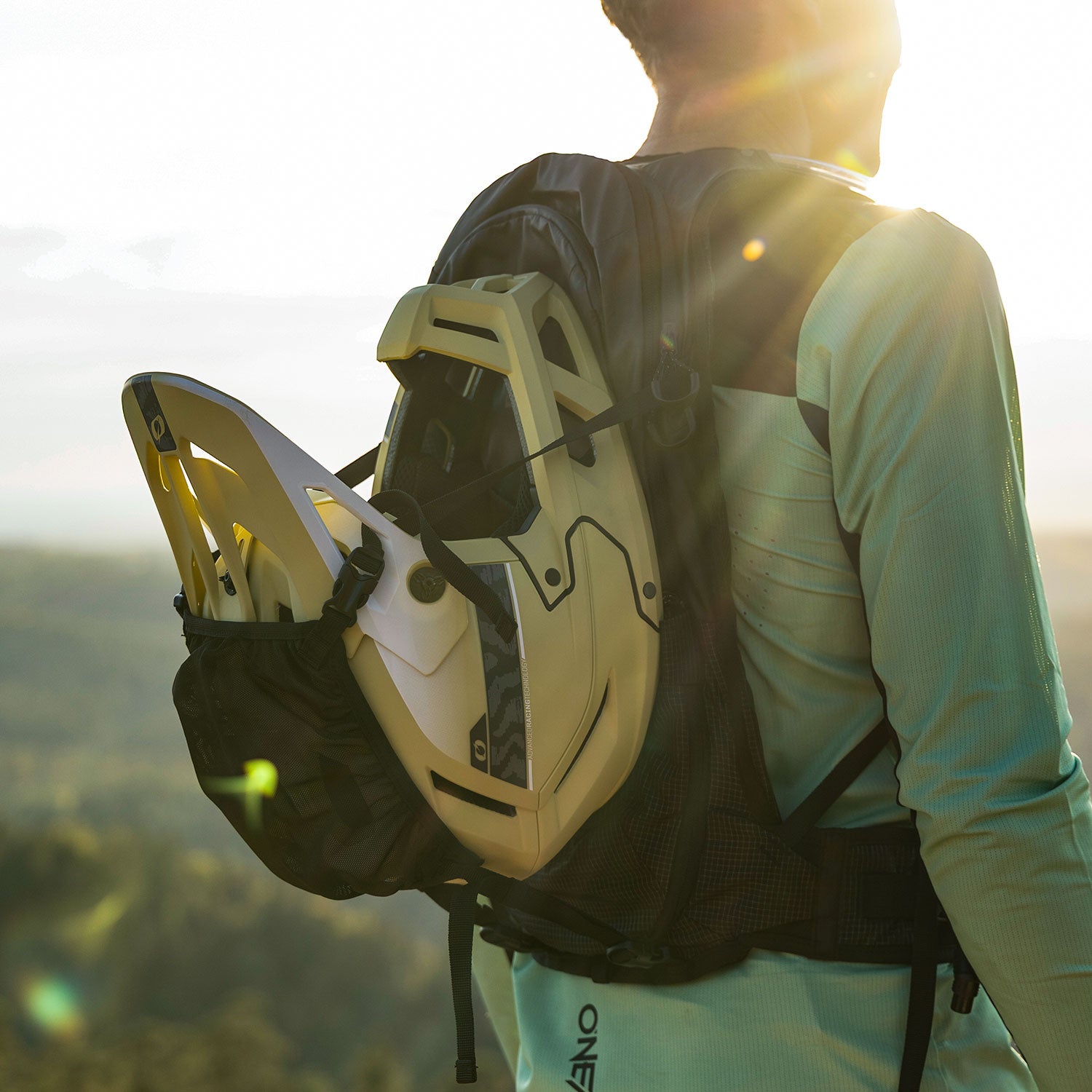 Eine Person mit einem hellgrünen Hemd und einem Rucksack, an dem ein gelber Mountainbike-Helm befestigt ist, steht im Freien im Sonnenlicht mit einer malerischen Landschaft im Hintergrund.