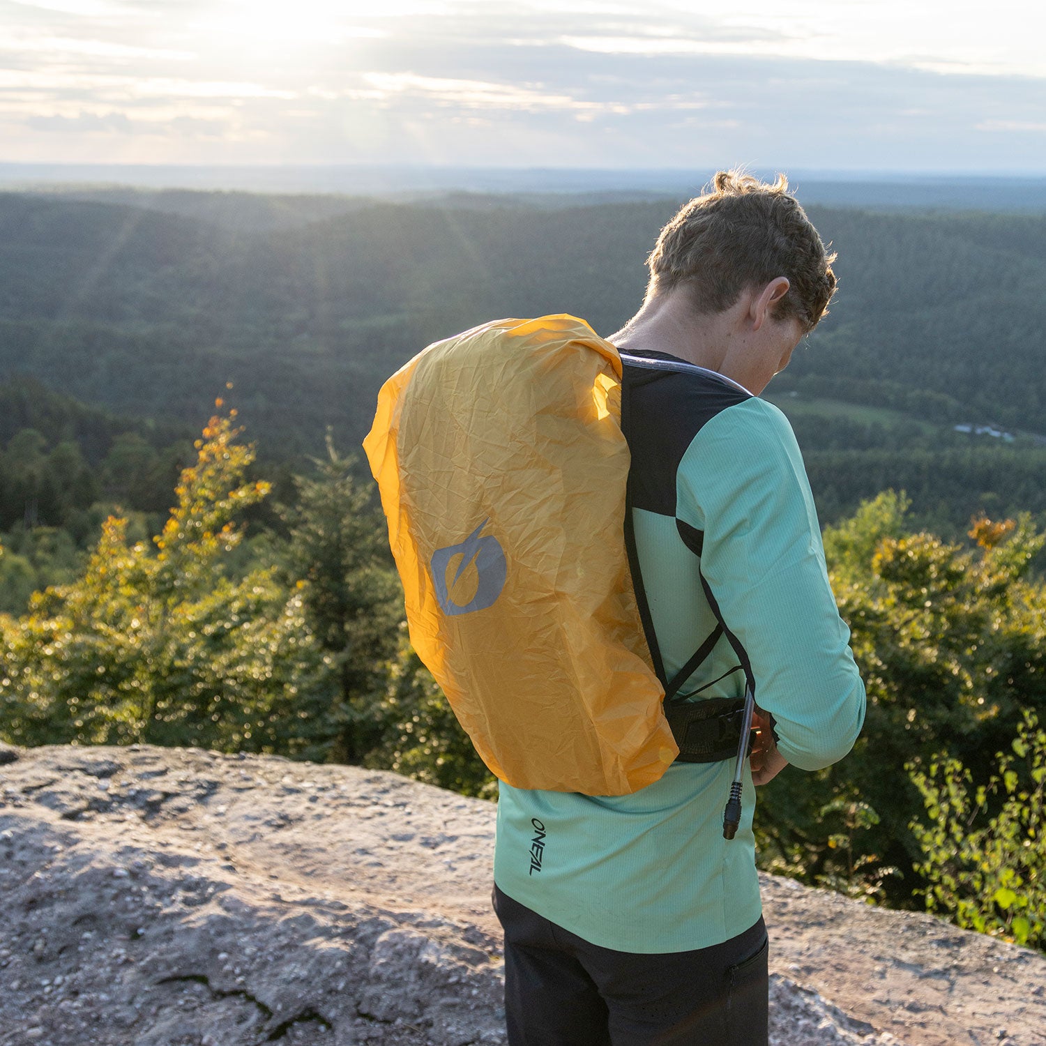 Eine Person in einer hellgrünen Jacke steht bei Sonnenuntergang auf einem Felsvorsprung mit Blick auf ein bewaldetes Tal, trägt eine gelbe Regenhülle auf ihrem Rucksack und verstellt einen Gurt.