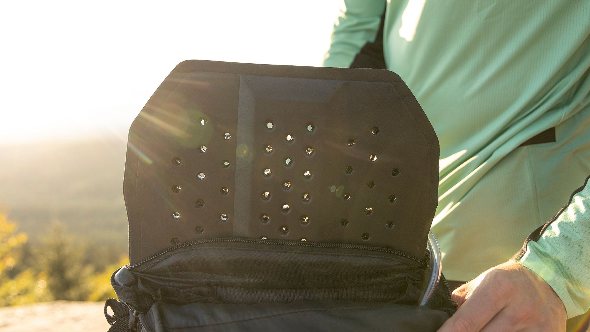 Eine Person in einem hellgrünen, langärmeligen Hemd öffnet einen Rucksack und enthüllt eine schwarze, belüftete Rückwand mit kreisförmigen Löchern. Im Hintergrund scheint das Sonnenlicht und schafft eine warme Atmosphäre im Freien.